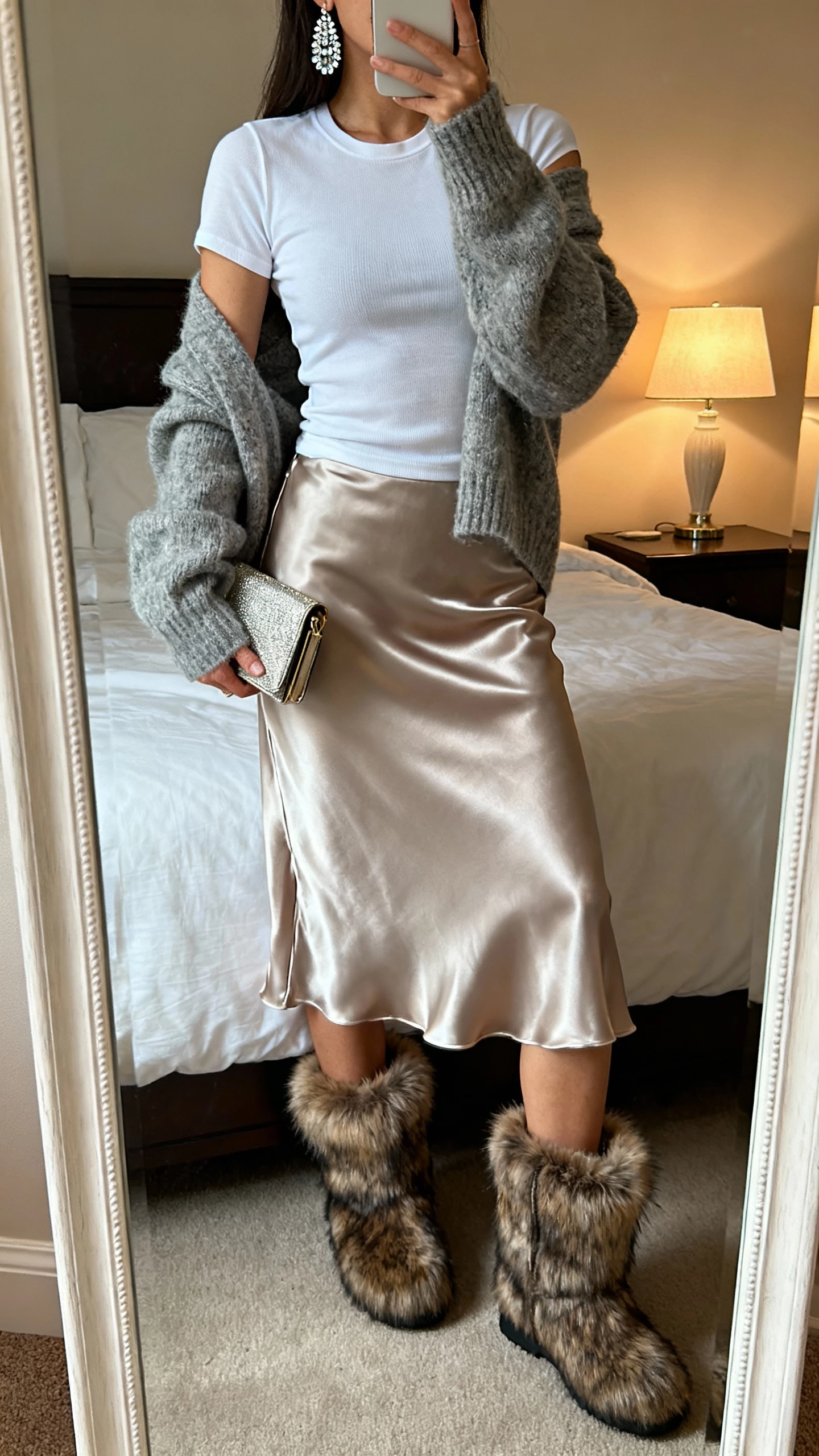 iPhone bedroom mirror selfie of a woman wearing faux-fur-lined winter booties with a satin slip skirt, cozy off-shoulder sweater layered over a fitted tee, sparkly earrings, and a small clutch, face not visible, soft evening lamp light, natural iPhone photo quality.