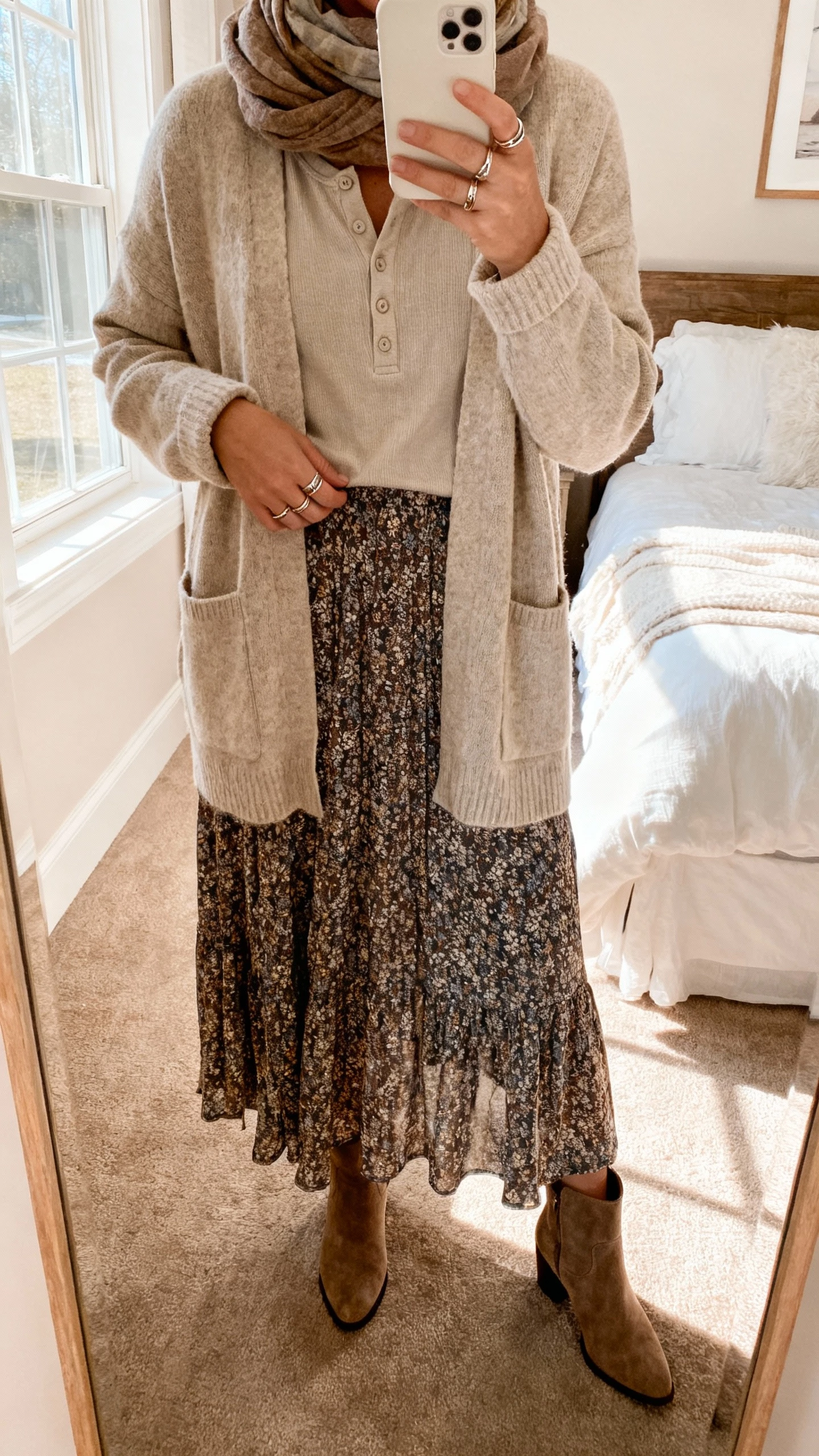 iPhone bedroom mirror selfie of a woman wearing layered knits and a long skirt: oatmeal cardigan over a soft henley, long flowy printed skirt, suede ankle boots, stacked rings and scarf, face not visible, warm window light, natural iPhone photo quality.