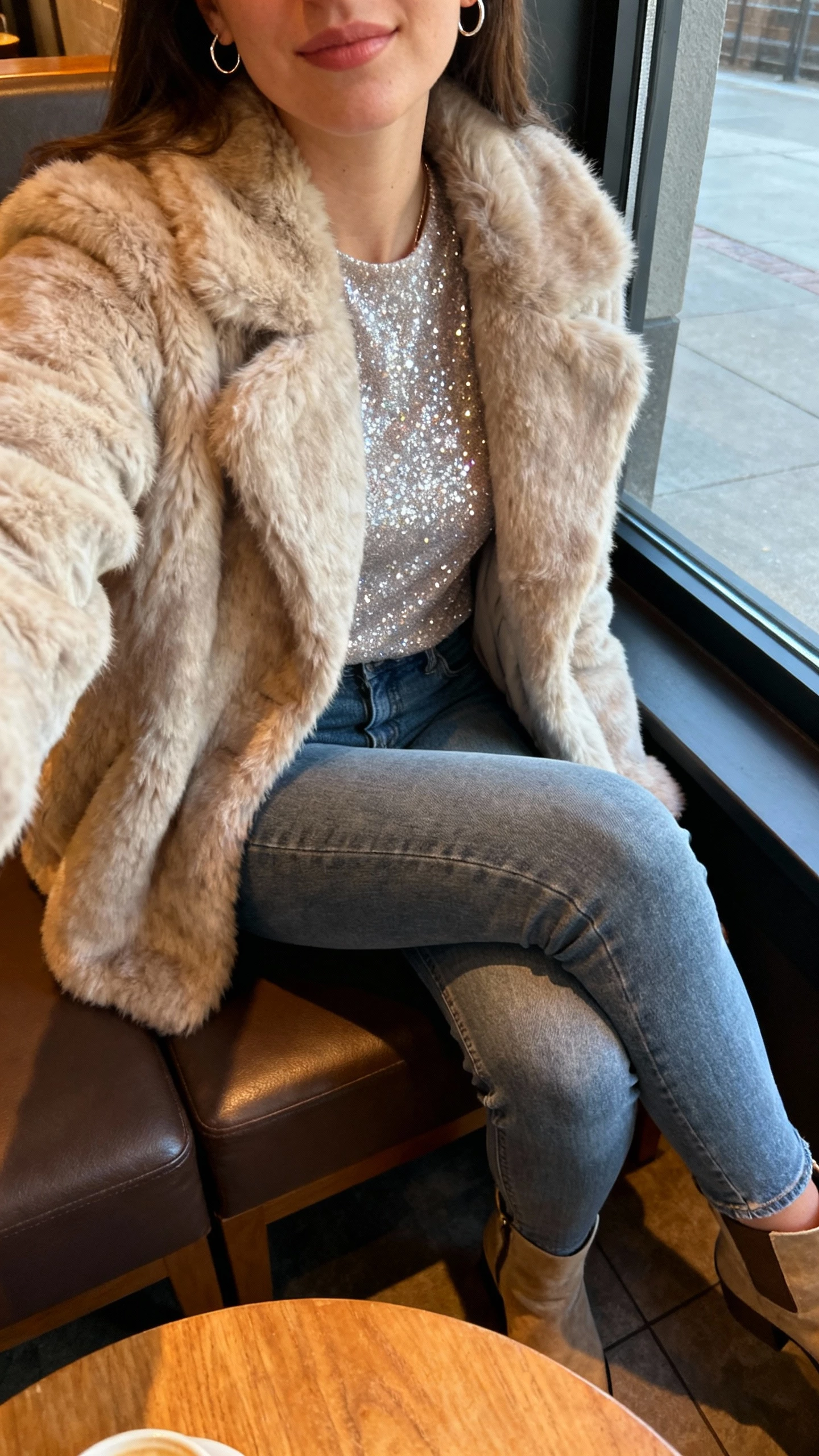 iPhone cafe seat selfie of a woman in a plush faux-fur coat over slim denim and a sparkle top, ankle boots and dainty hoops, face not visible, cropped at shoulders, warm cafe lighting near window, natural iPhone photo quality.