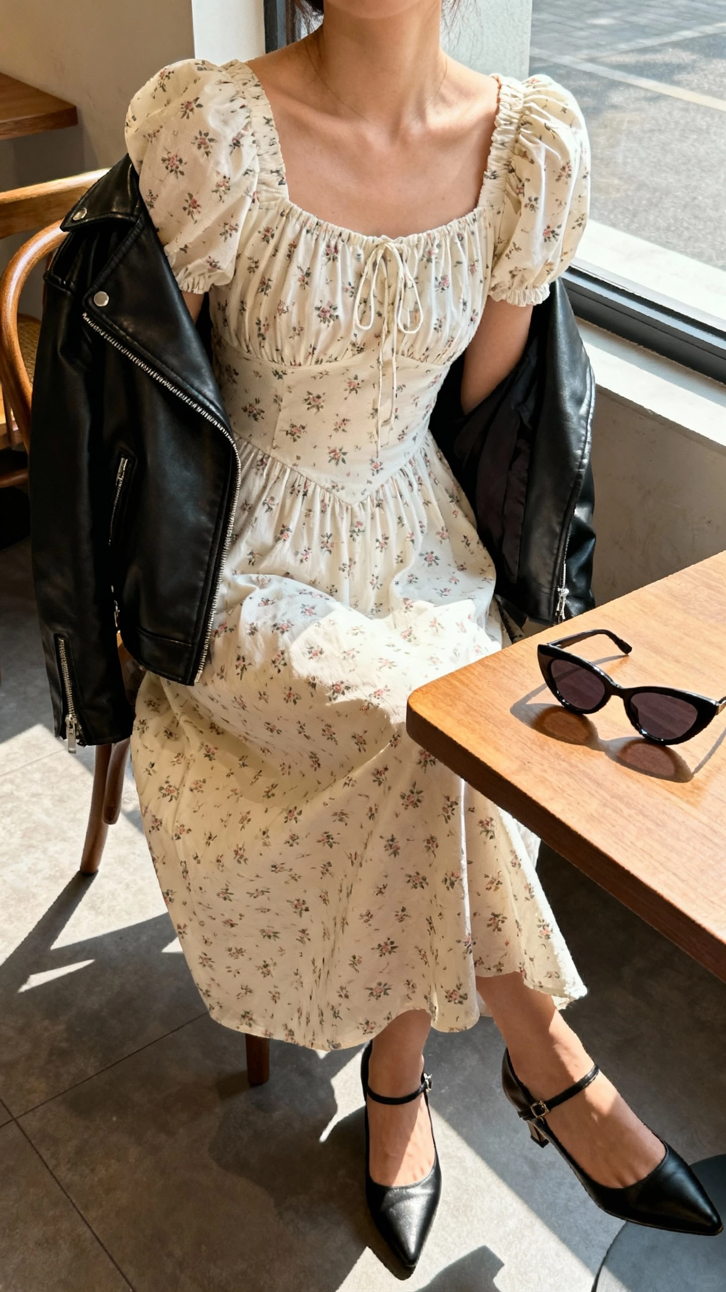 iPhone cafe selfie cropped at neck showing a woman wearing a vintage-inspired tea dress with tiny florals, puff sleeves, modern leather moto jacket draped over shoulders, kitten heels, and cat-eye sunglasses on table, face not visible, window light, natural iPhone photo quality