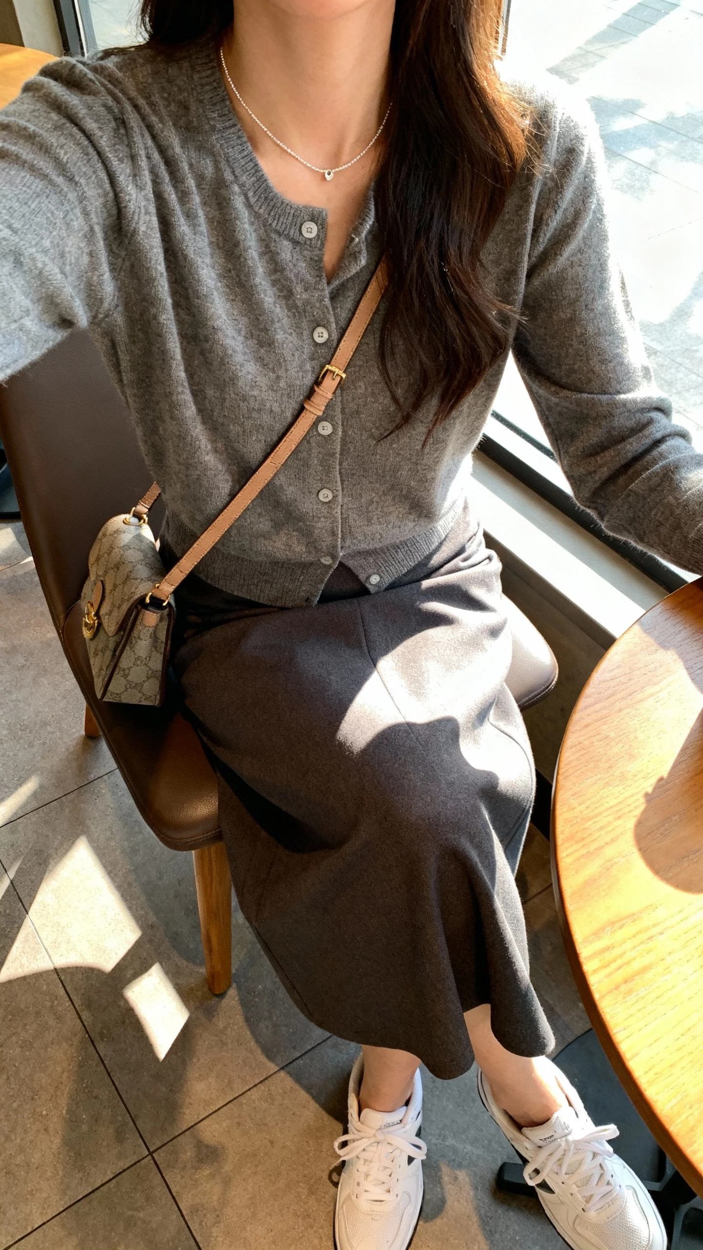 iPhone cafe table selfie cropped at neck showing a woman in a polished cardigan over a midi skirt with clean white sneakers, dainty necklace, crossbody bag on chair, face not visible, window light in coffee shop, natural iPhone photo quality.