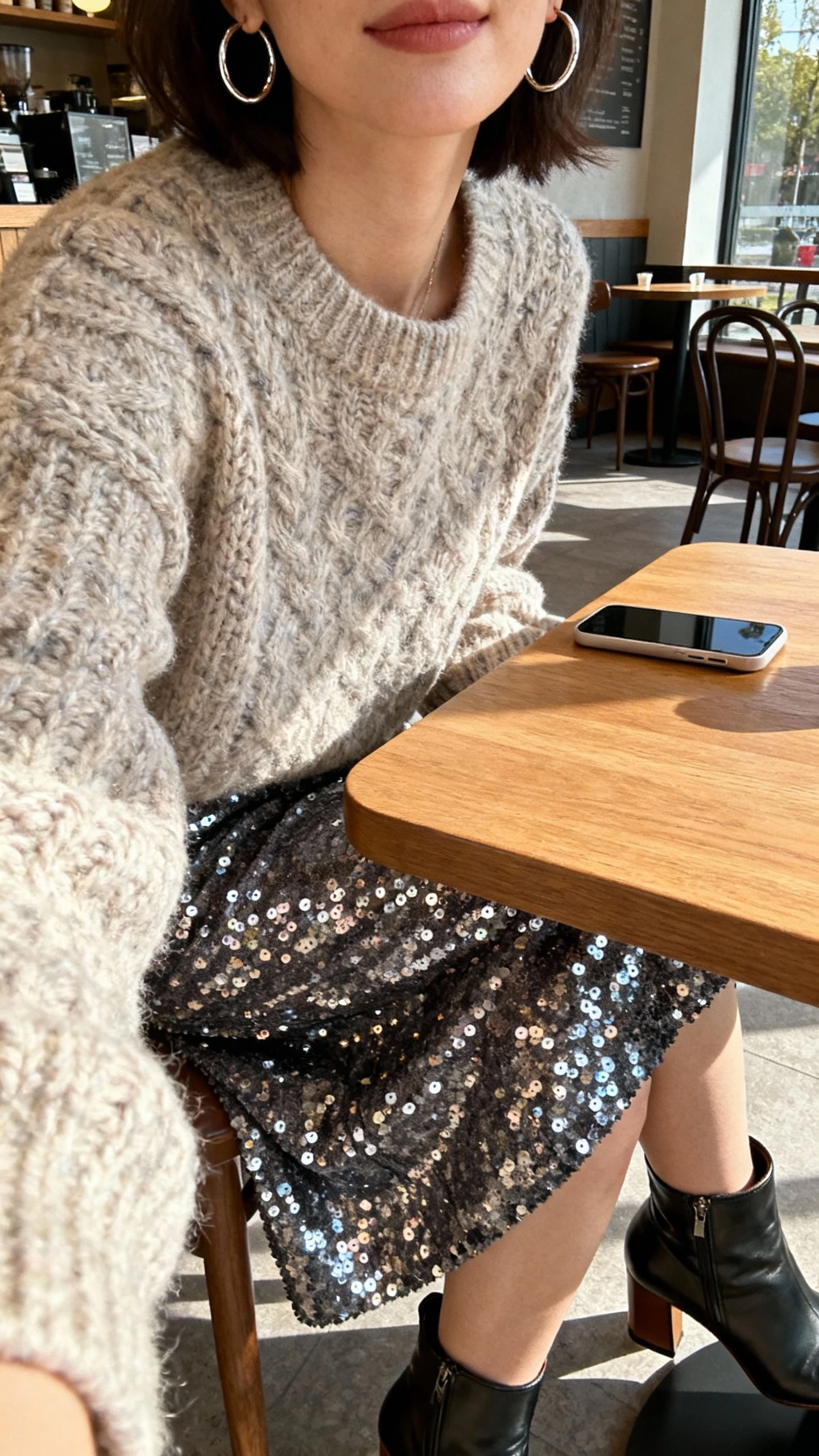 iPhone cafe table selfie cropped at neck showing a woman wearing a sequin skirt with a chunky sweater, hoop earrings, and heeled ankle boots, face not visible, cozy cafe setting with natural daylight, natural iPhone photo quality.