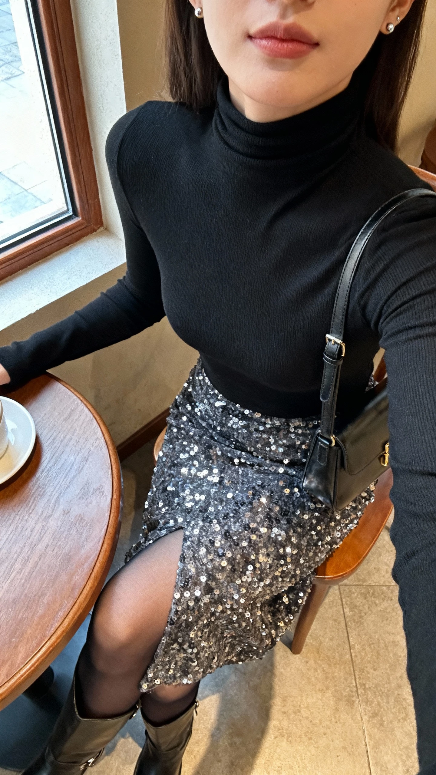 iPhone cafe table selfie cropped at neck showing a woman in a black fitted turtleneck layered under a sequin midi skirt, sheer tights, ankle boots, small shoulder bag, simple studs, face not visible, warm indoor lighting by window, natural iPhone photo quality