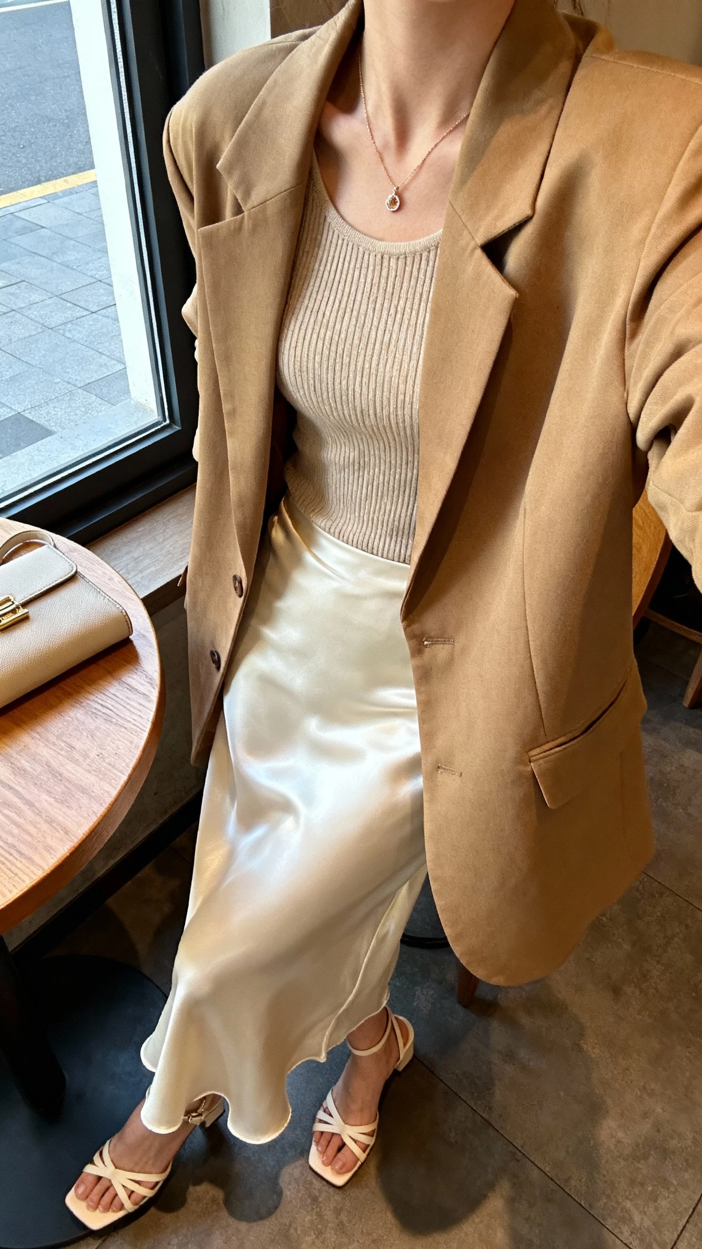 iPhone cafe table selfie cropped at neck showing a woman in a cream satin slip skirt, oversized tan blazer over a fitted knit top, strappy heeled sandals, dainty pendant necklace, clutch on table, face not visible, warm cafe light by window, natural iPhone photo.