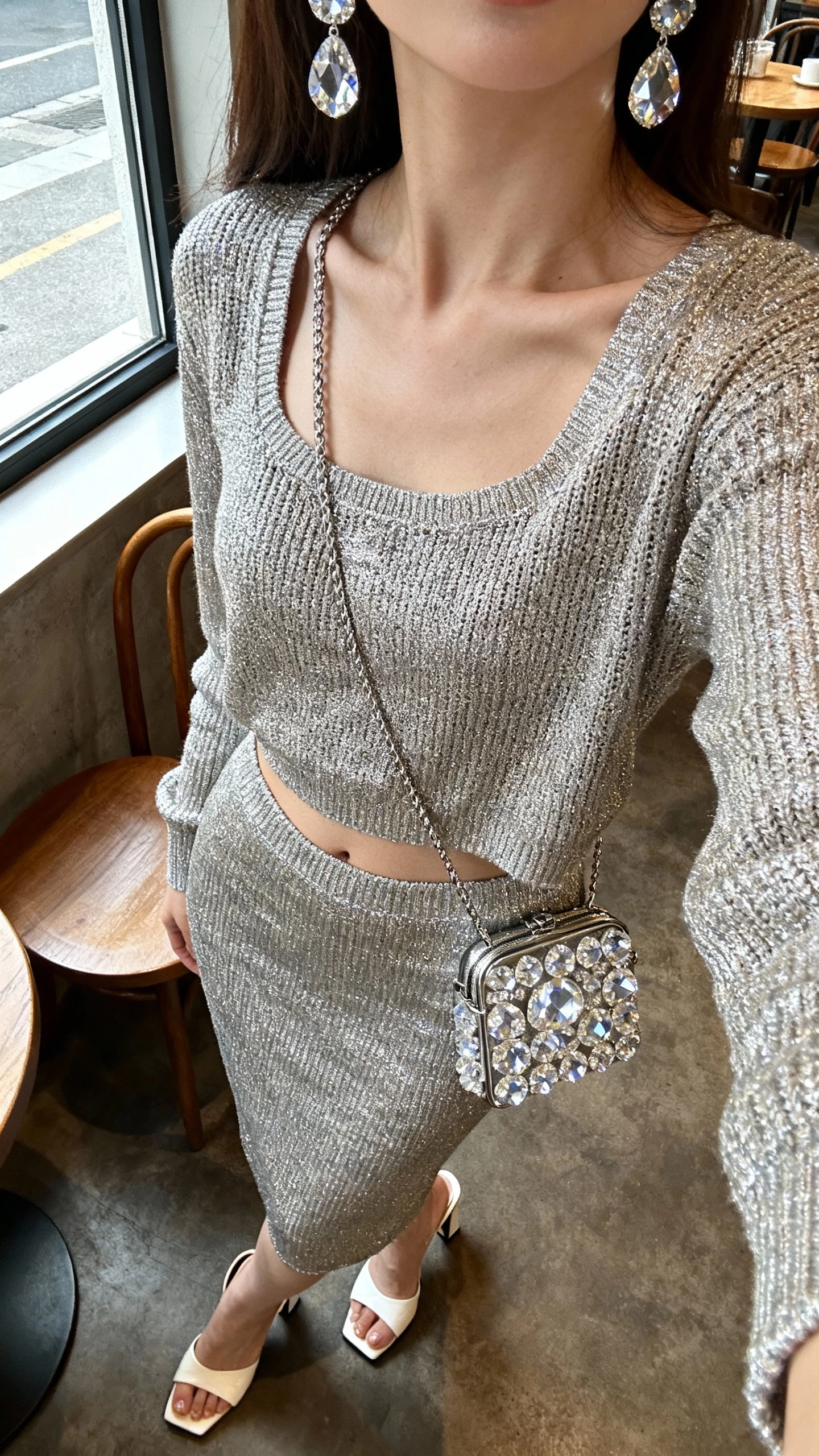 iPhone cafe table selfie cropped at neck showing a woman wearing a metallic knit set (matching top and skirt) with crystal earrings and a crystal-embellished mini bag, heeled mules, face not visible, cozy cafe window light, natural iPhone aesthetic.