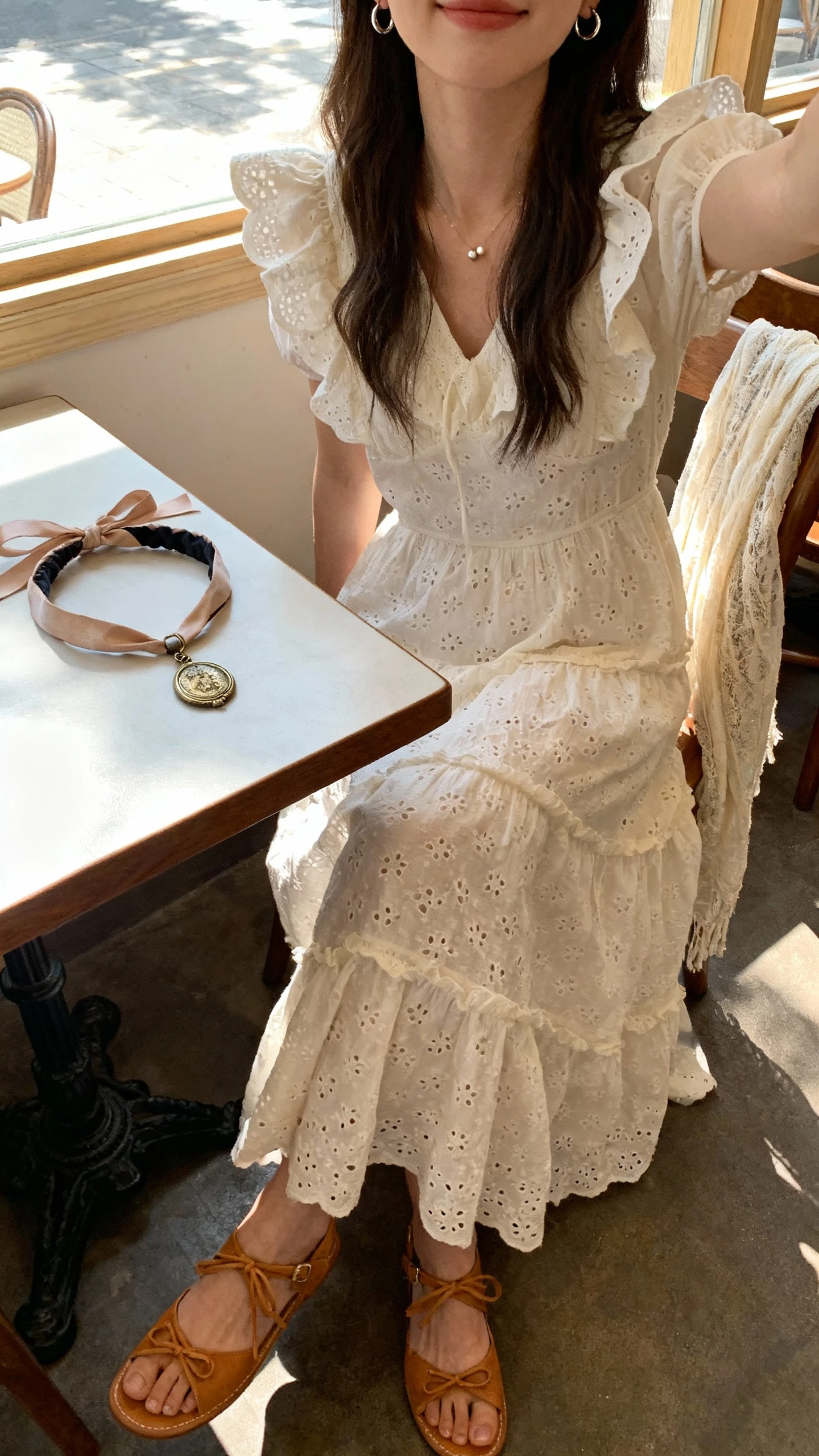 iPhone cafe table selfie cropped at neck showing a woman in an eyelet tiered gown with flutter sleeves, lace-up sandals in tan, ribboned headband set on the table, vintage locket, tiny hoop earrings, lightweight shawl draped over chair, face not visible, sunlit cafe window, natural iPhone aesthetic.