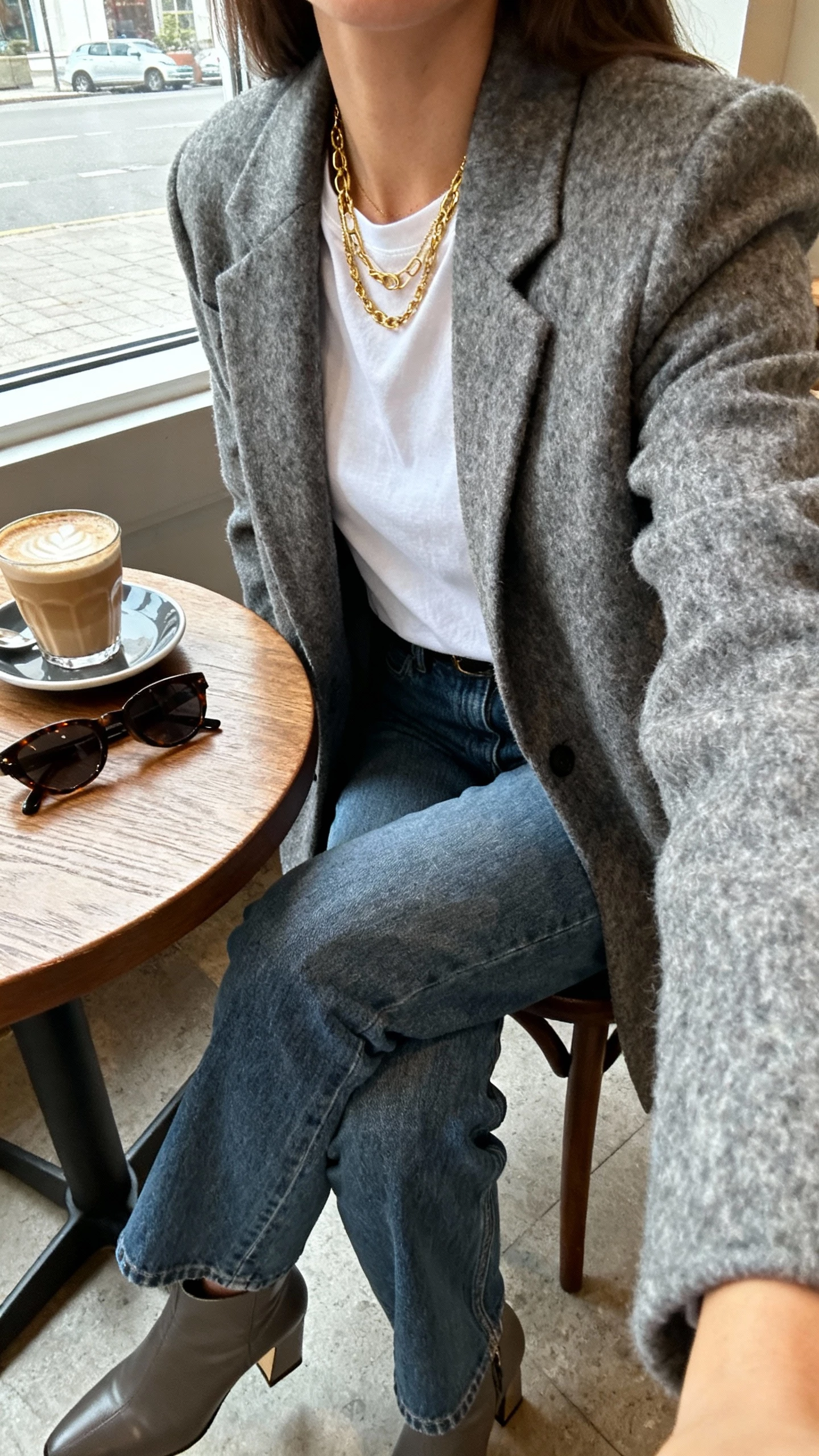 iPhone cafe table selfie cropped at shoulders showing a woman wearing a gray wool blazer over a white tee with straight-leg blue jeans, heeled ankle boots, layered gold necklaces, latte and sunglasses on table, face not visible, cozy coffee shop window light, natural iPhone photo quality