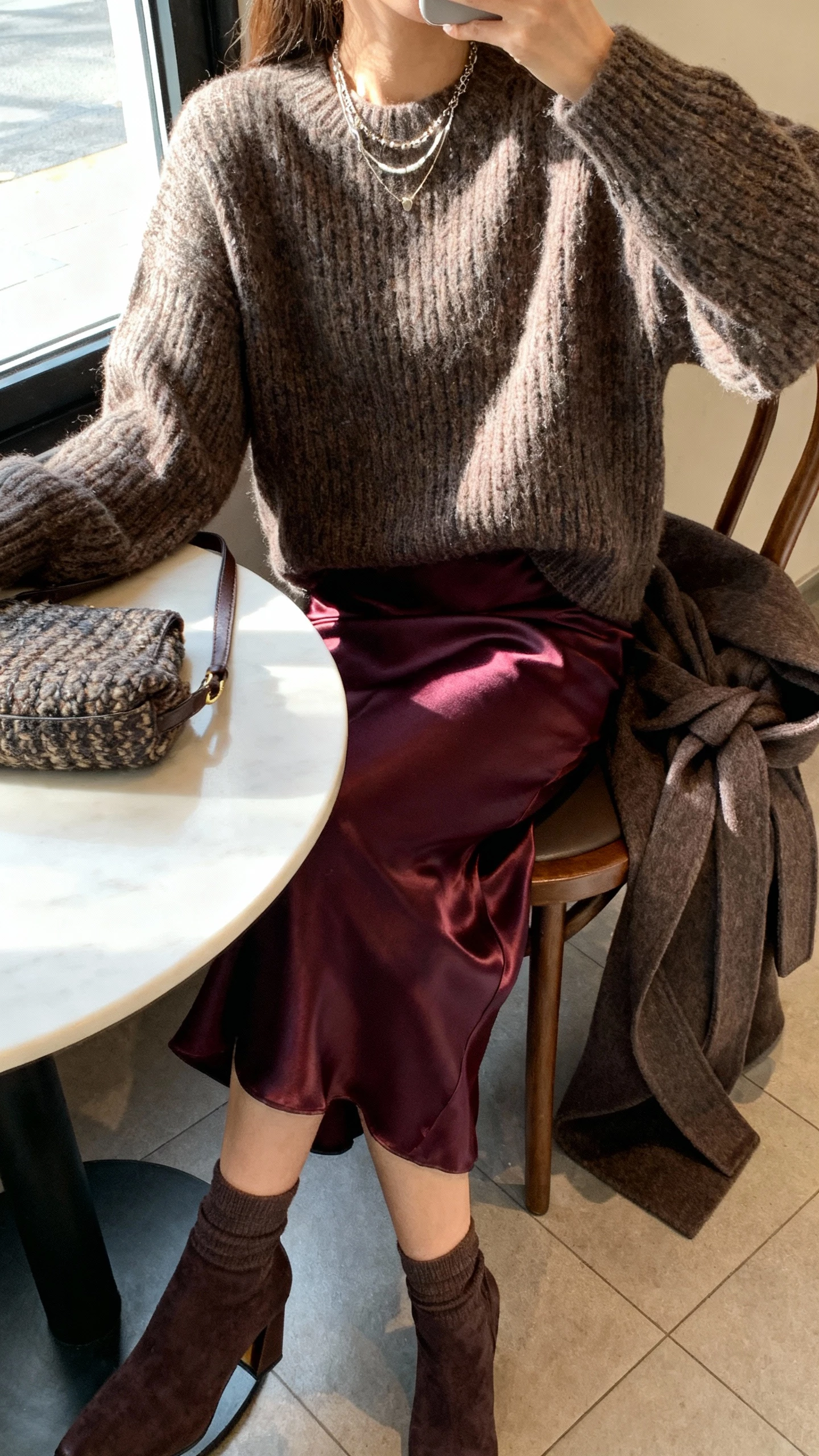 iPhone cafe table selfie cropped at shoulders showing a woman wearing a deep jewel‑tone satin slip skirt, oversized fisherman sweater, heeled sock boots, classic wool wrap coat draped over chair, textured shoulder bag, layered necklaces; face not visible, window light, natural iPhone photo quality.