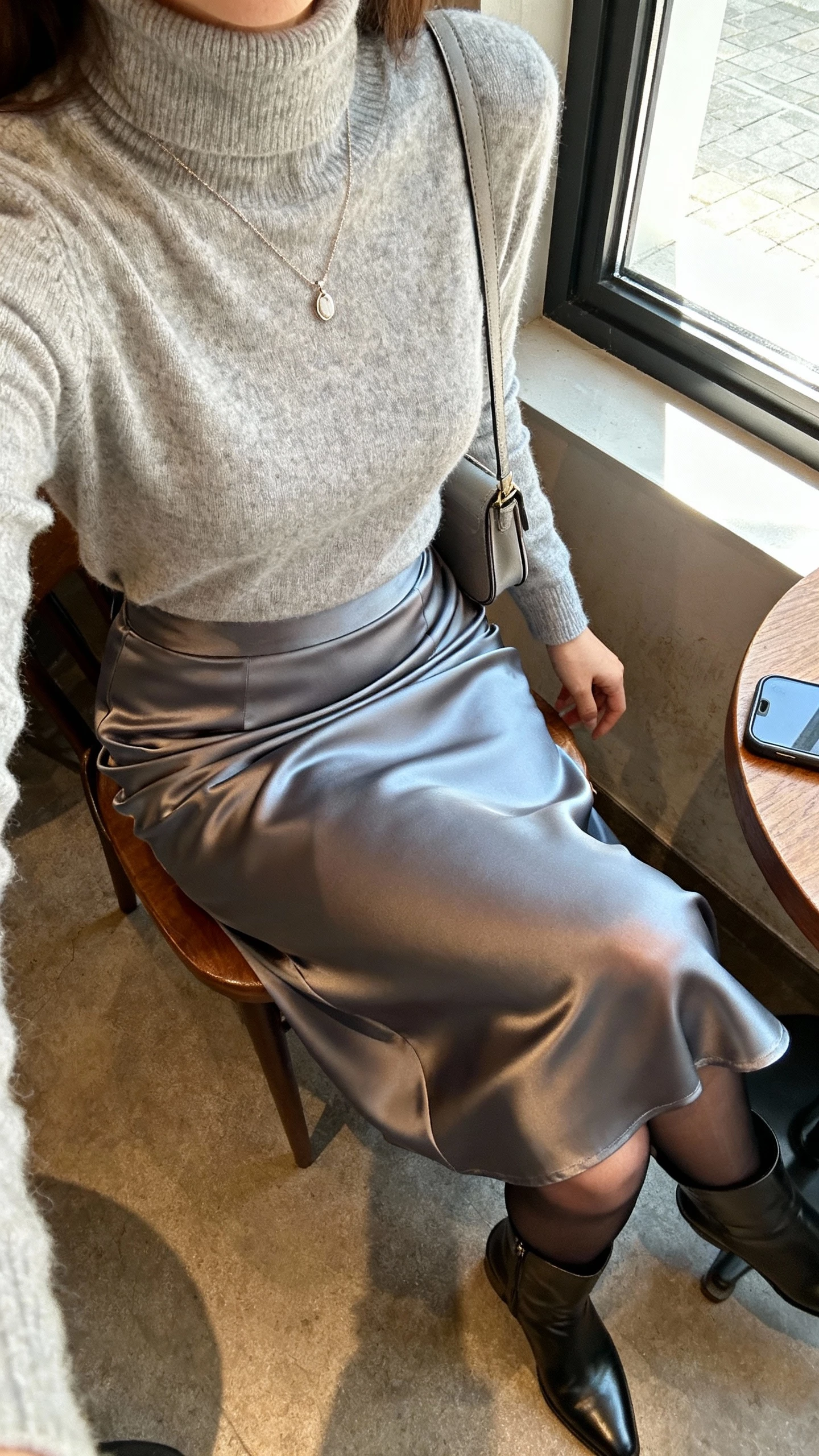 iPhone cafe table selfie cropped at shoulders showing a woman wearing a cashmere turtleneck tucked into a bias-cut satin midi skirt, sheer tights, sleek ankle boots, delicate pendant necklace, and a small shoulder bag, face not visible, morning window light, natural iPhone photo quality.