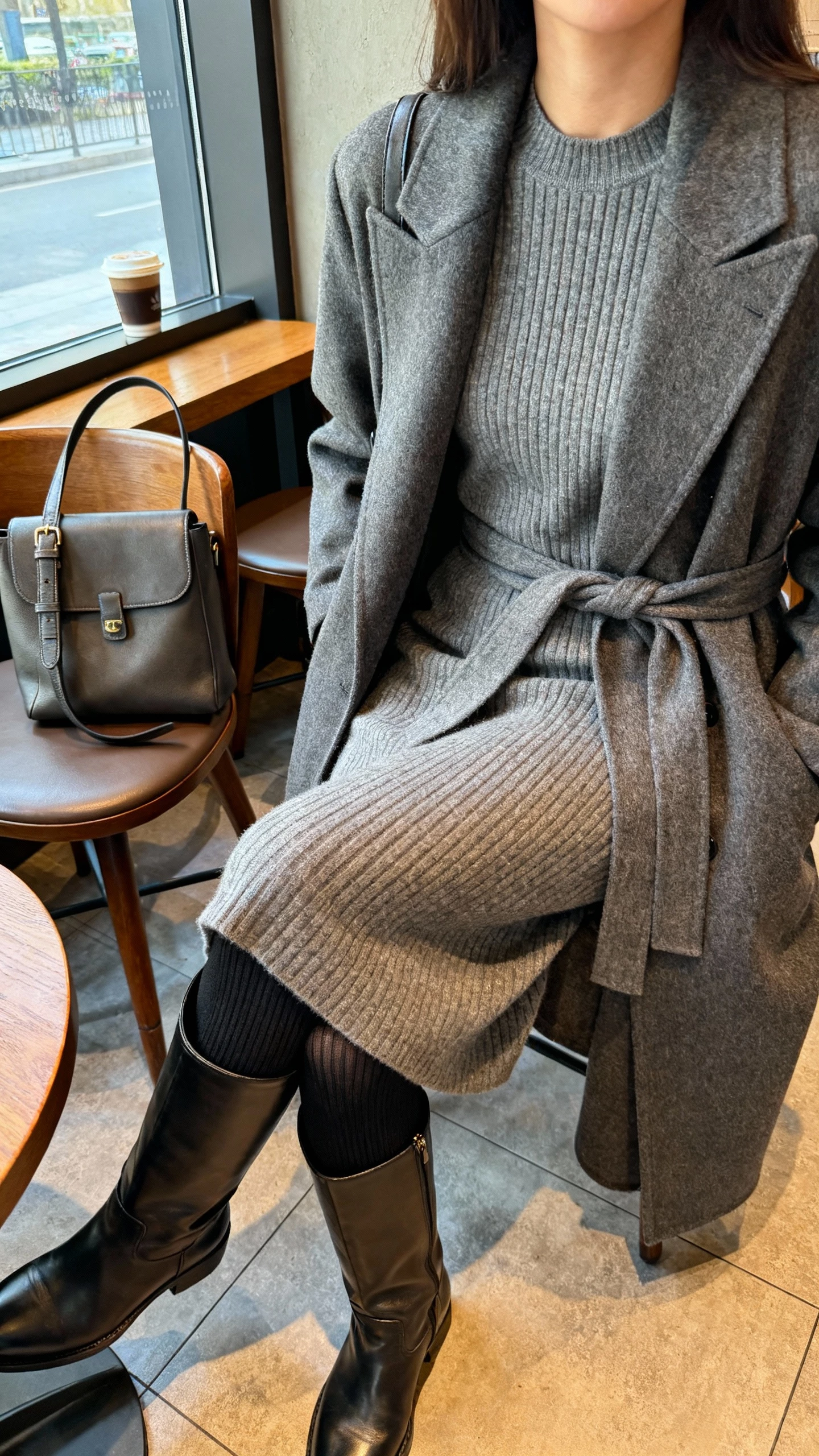 iPhone cafe table selfie cropped at shoulders showing a woman wearing a heather grey ribbed midi knit dress with a tailored wrap coat, leather knee-high boots, black thermal tights, and a structured shoulder bag set on the chair; face not visible, cozy coffee shop window light, natural iPhone photo quality.