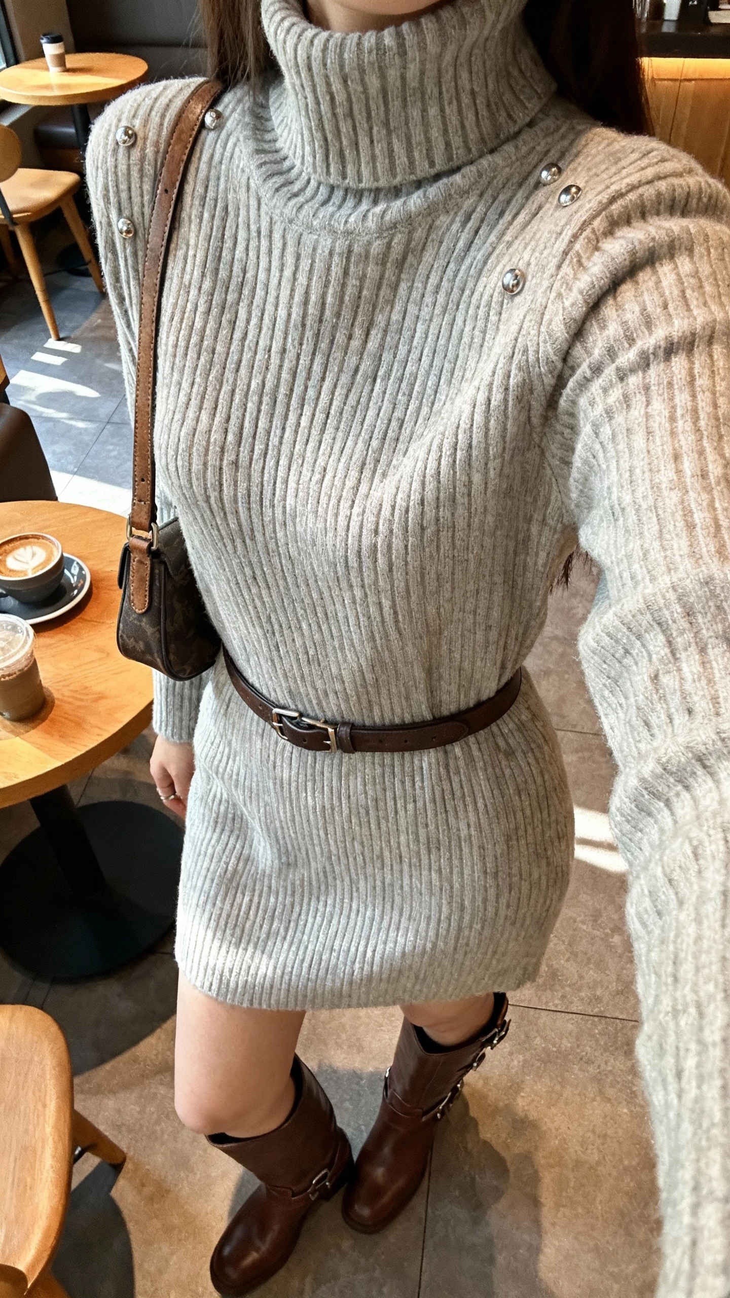 iPhone cafe table selfie cropped at shoulders showing a woman in a ribbed turtleneck sweater dress with a belt, knee-high leather boots, small shoulder bag, simple studs, face not visible, cozy coffee shop setting with afternoon light, natural iPhone quality.