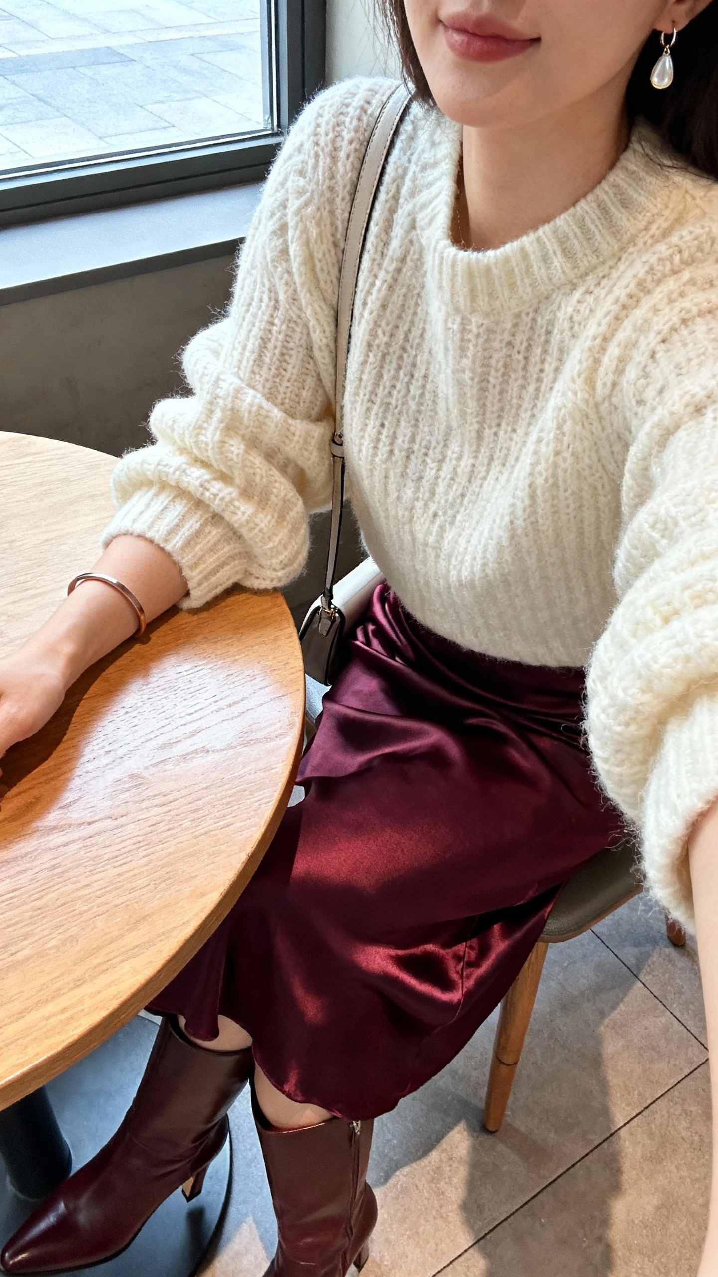 iPhone cafe table selfie cropped at shoulders showing a woman in a deep berry satin slip skirt, chunky cream sweater, tall heeled leather boots, simple drop earrings with a thin cuff bracelet, slim shoulder bag; face not visible, soft window light, natural iPhone photo quality.