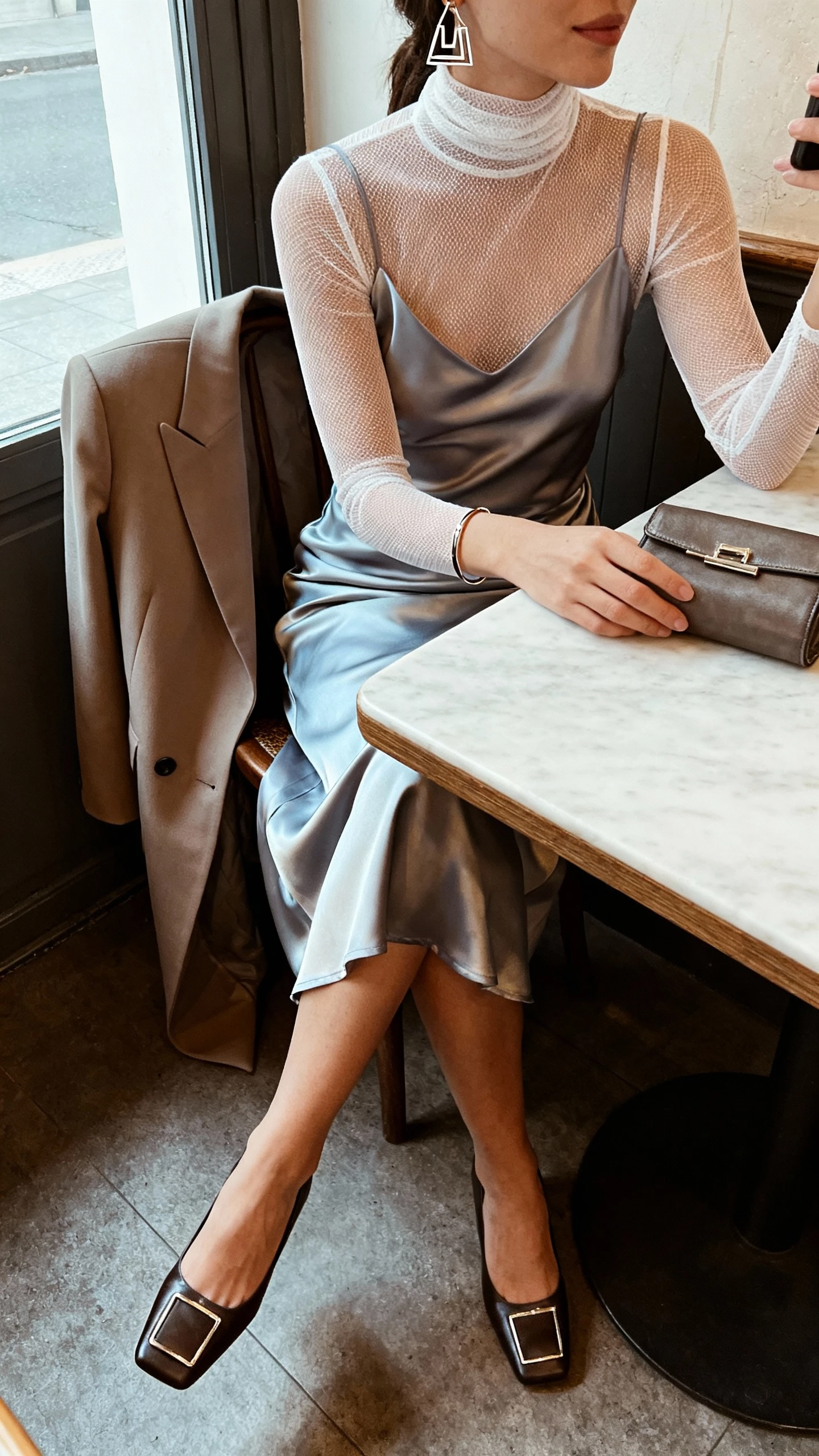 iPhone cafe table selfie cropped at shoulders showing a woman in a bias-cut satin slip dress layered over a sheer mesh turtleneck, square-toe heels, geometric earrings, a slim bracelet, a structured clutch, and a long tailored coat on the chair; face not visible, morning window light, natural iPhone aesthetic.