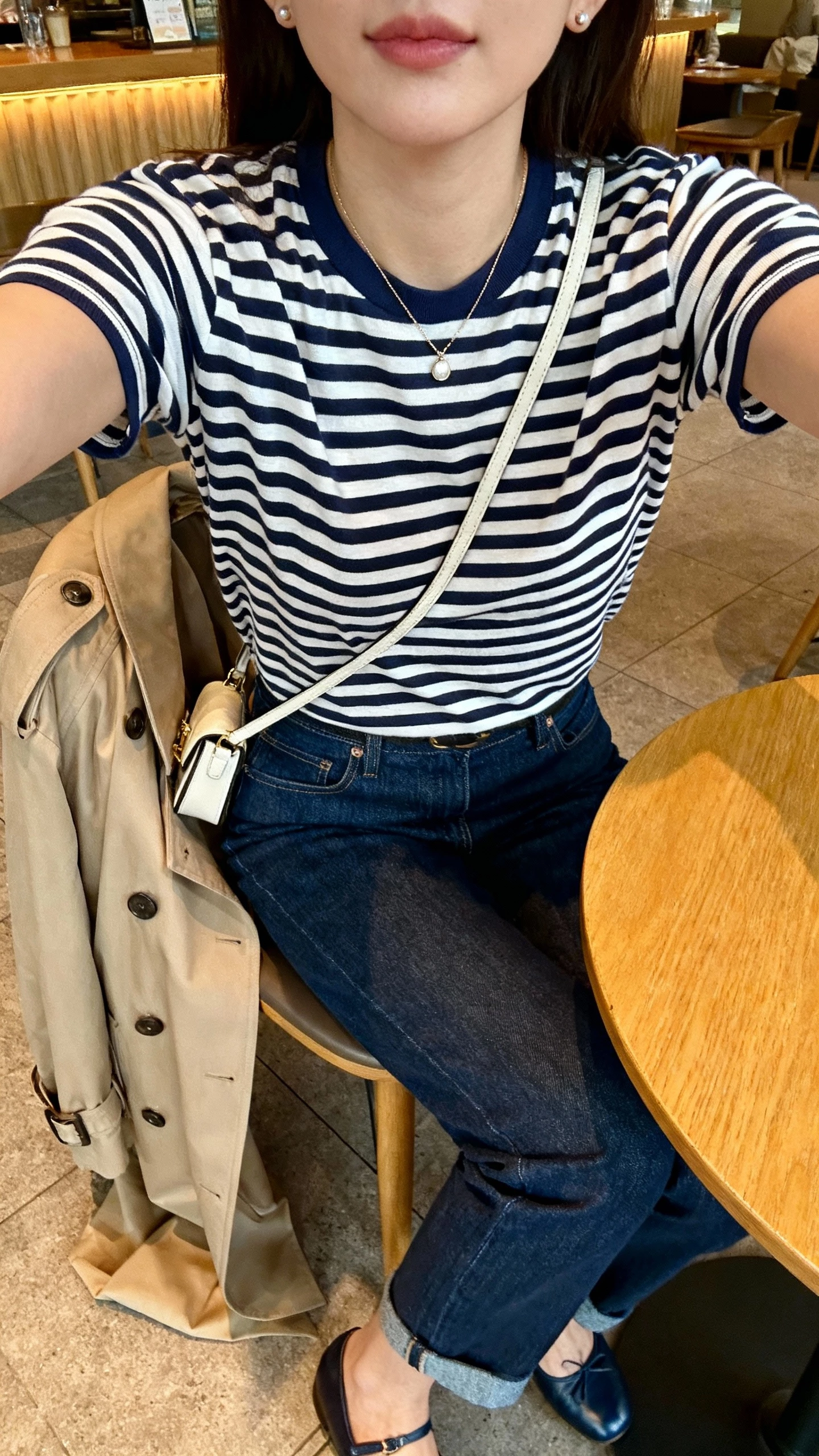 iPhone cafe table selfie cropped at shoulders showing a woman in a navy-and-white Breton striped tee with dark blue slightly cropped straight-leg jeans, ballet flats, trench coat over chair, delicate pendant, small studs, and a mini crossbody; warm cafe lighting, face not visible, natural iPhone photo.