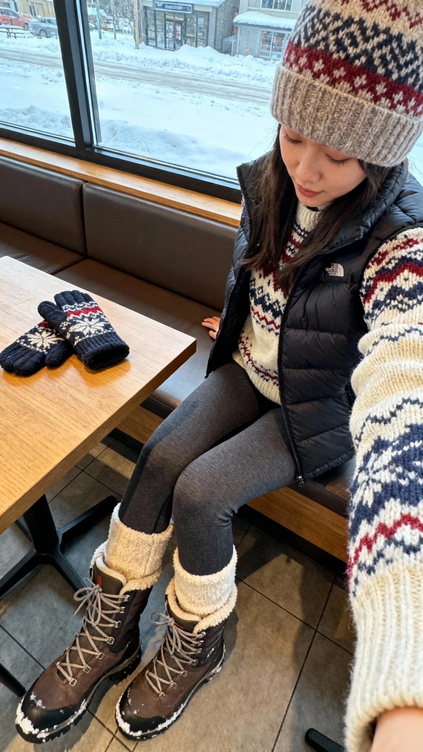 iPhone cafe table selfie (downward angle) of a woman in a Fair Isle sweater, fleece-lined leggings, puffer vest, snow-ready lace-up boots, knit beanie, mittens on the table, face not visible, cozy cafe window seat with snowy view, natural daylight, iPhone photo quality.