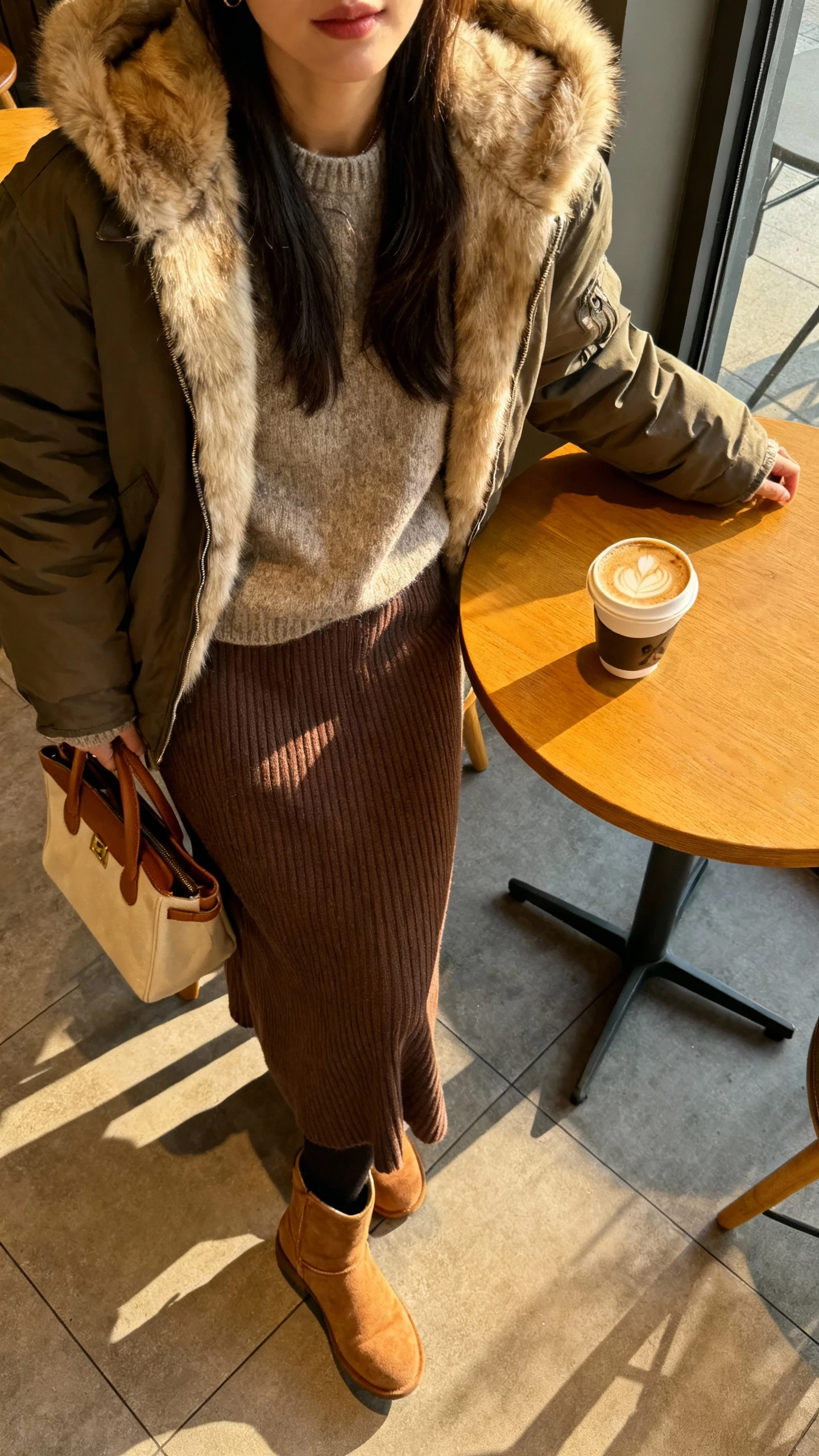 iPhone cafe table selfie (downward angle) of a woman wearing a fur-lined hooded jacket, ribbed knit midi skirt, cozy sweater, ankle boots, tote with coffee cup, face not visible, warm cafe morning light, natural iPhone photo quality.
