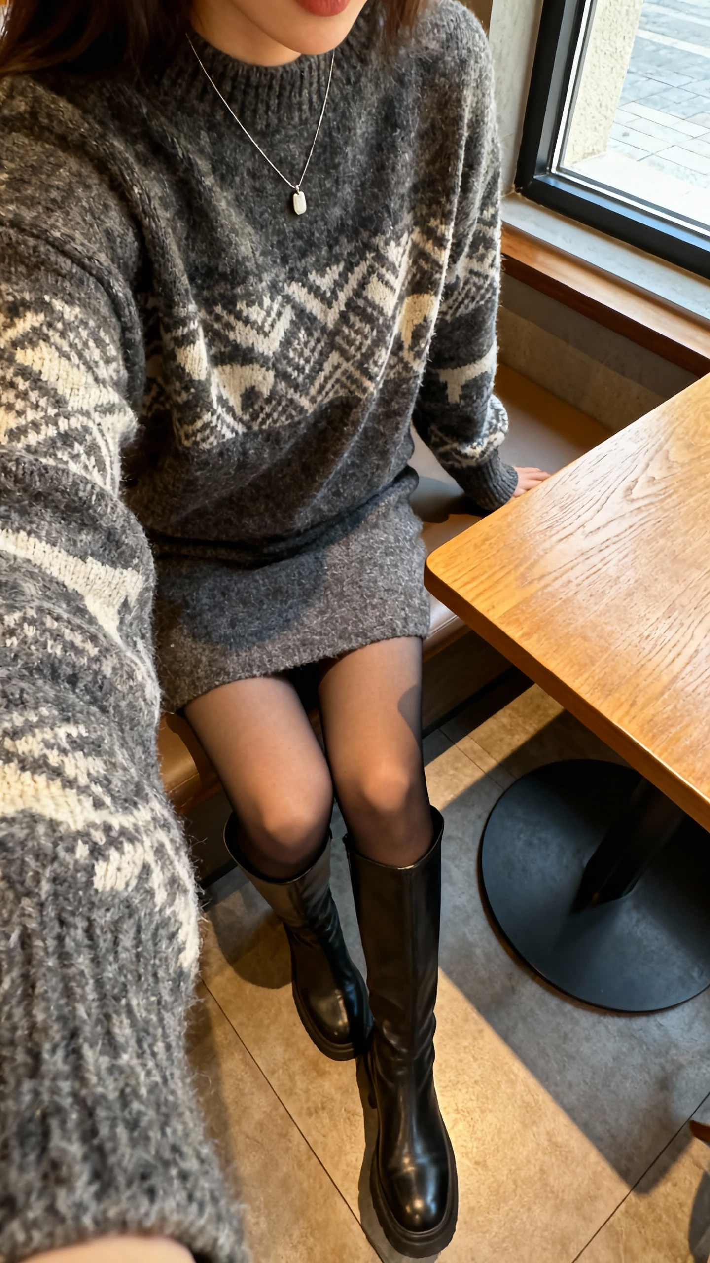 iPhone cafe table selfie, downward angle, of a woman wearing a statement sweater dress with tall knee-high boots, opaque tights, simple pendant necklace, face not visible, warm cafe lighting by window, natural iPhone quality.