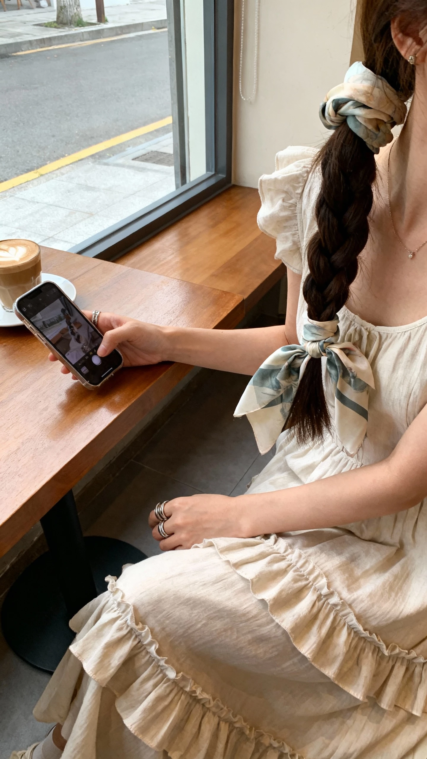 iPhone cafe table selfie from side angle of a woman with a boho pony wrapped in a braid and a silk scarf, wearing a flounced midi dress and stacked rings, face not visible, cozy coffee shop window light, natural iPhone photo quality.