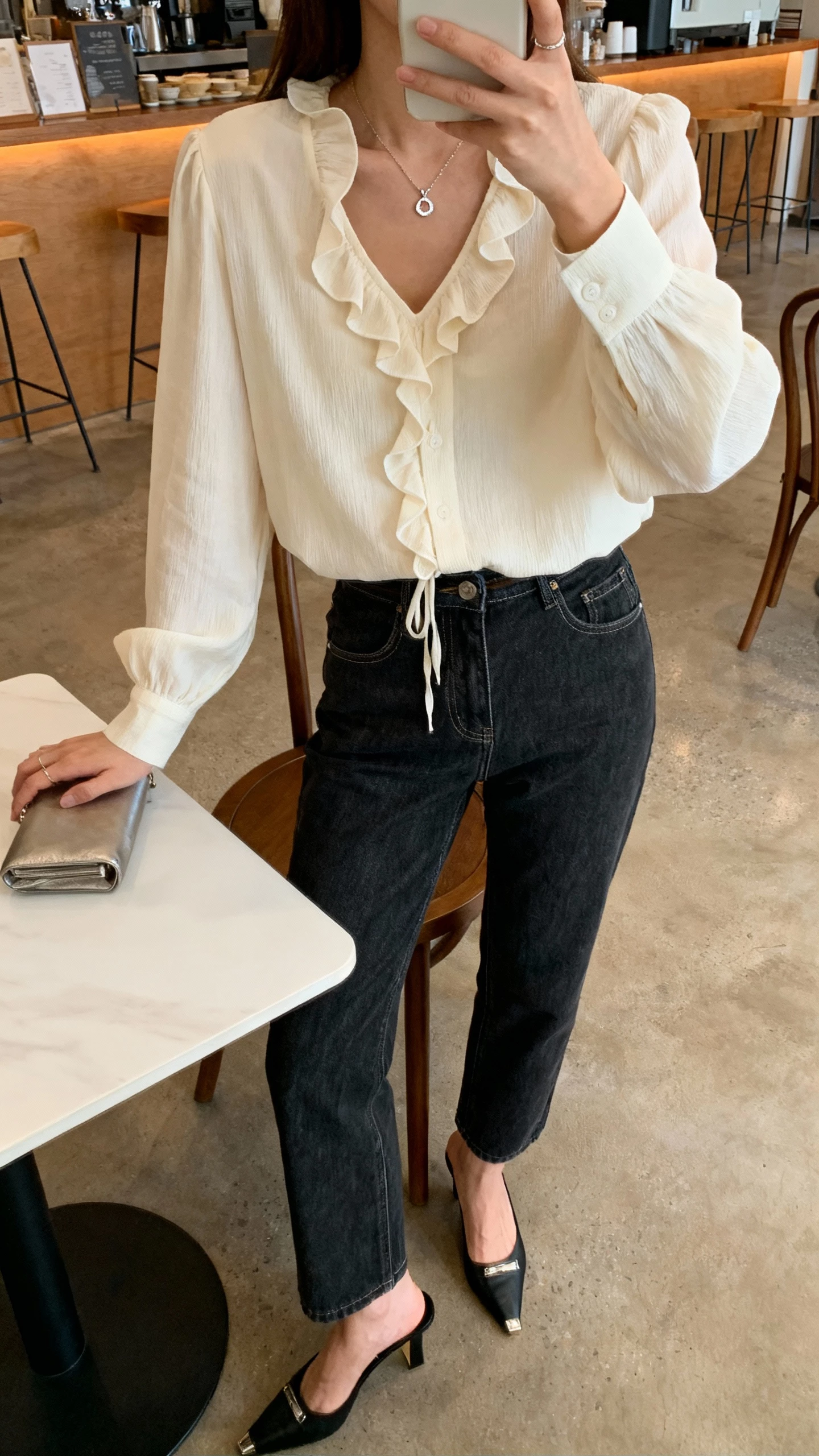 iPhone cafe table selfie of a woman wearing a romantic cream blouse with subtle ruffles tucked into dark-wash denim, heeled mules, dainty pendant, small clutch, face not visible, warm cafe lighting, natural iPhone photo.