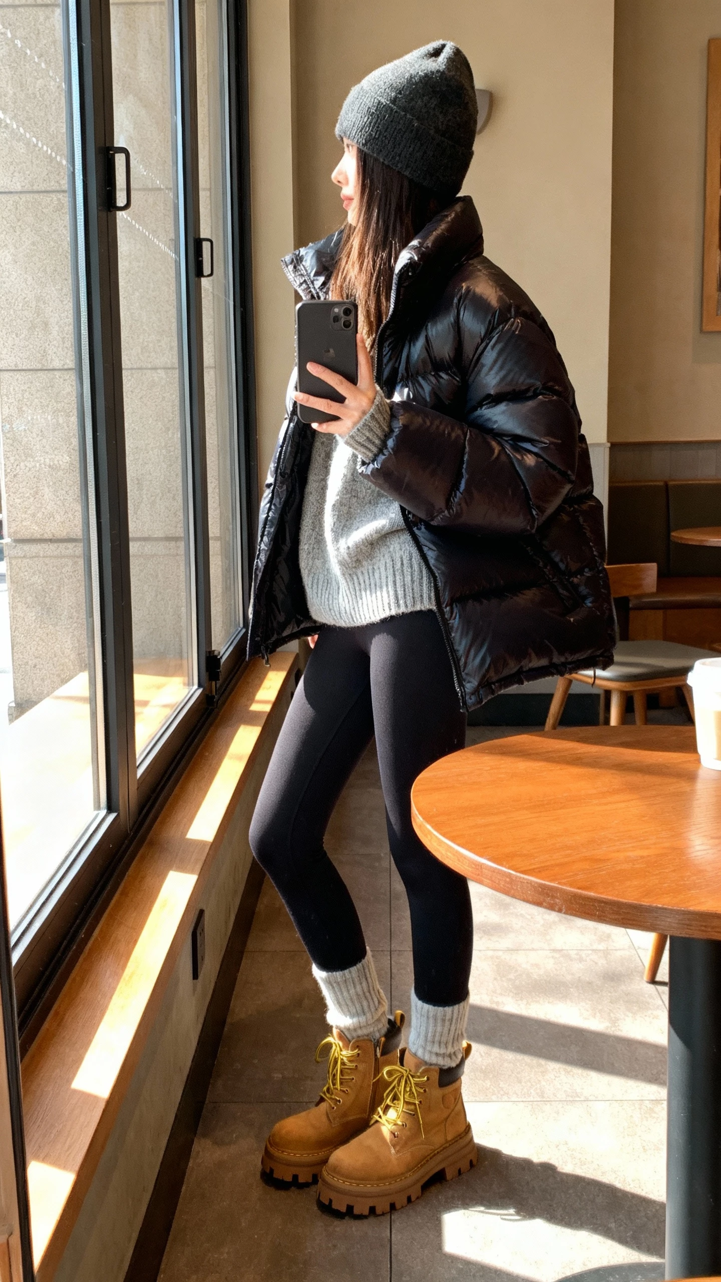 iPhone cafe table selfie of a woman wearing a sleek puffer over a cozy knit with sleek leggings, lug-sole boots and a beanie, face not visible, side angle, morning light by window, natural iPhone photo quality.