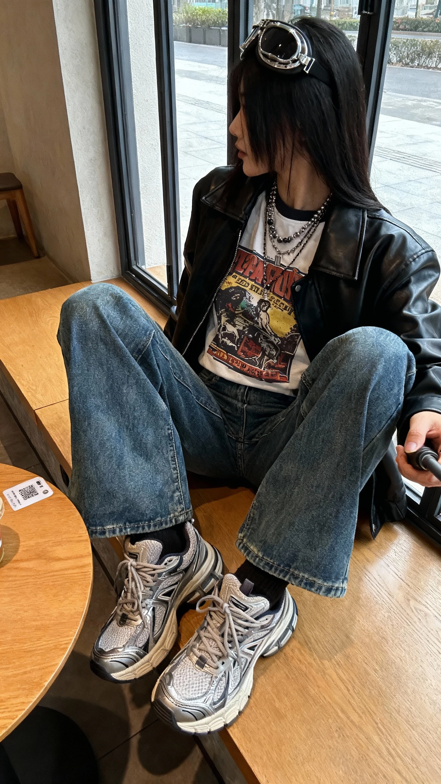iPhone cafe table selfie, side angle, of a woman wearing a vintage graphic tee, black leather jacket, indigo bootcut jeans, chunky sneakers, layered necklaces, aviator-style sunglasses on head; face not visible, window light, natural iPhone photo quality.