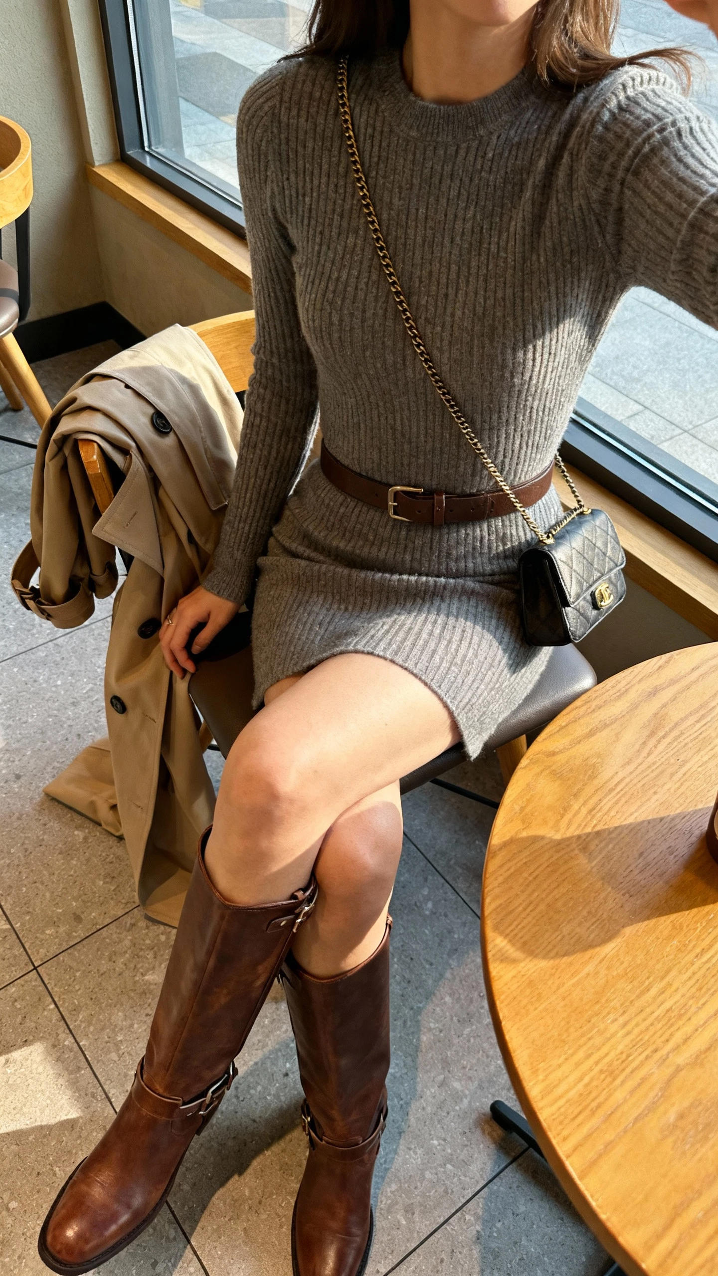 iPhone cafe table selfie, slightly top-down, showing a woman in a fitted sweater dress with tall knee-high leather boots, belt at waist, trench draped over chair, small crossbody bag, face not visible, warm indoor light by window, natural iPhone photo quality