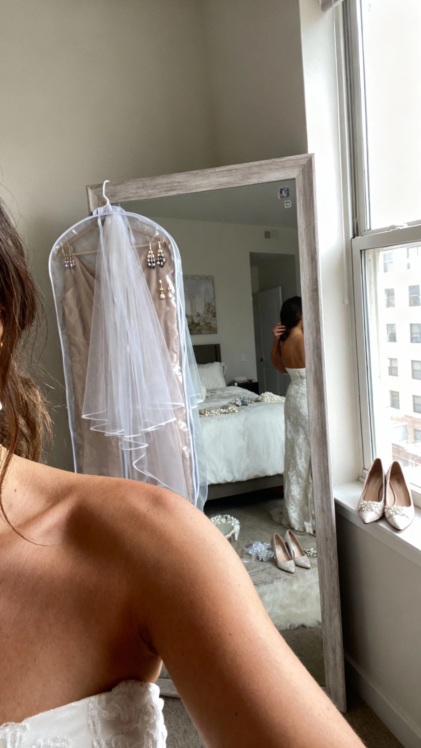 iPhone casual selfie cropped at shoulders of a woman holding a garment bag and trying on different wedding accessories (veil, earrings, shoes) next to a full-length mirror, face not visible, apartment bedroom with natural window light, natural iPhone photo quality.