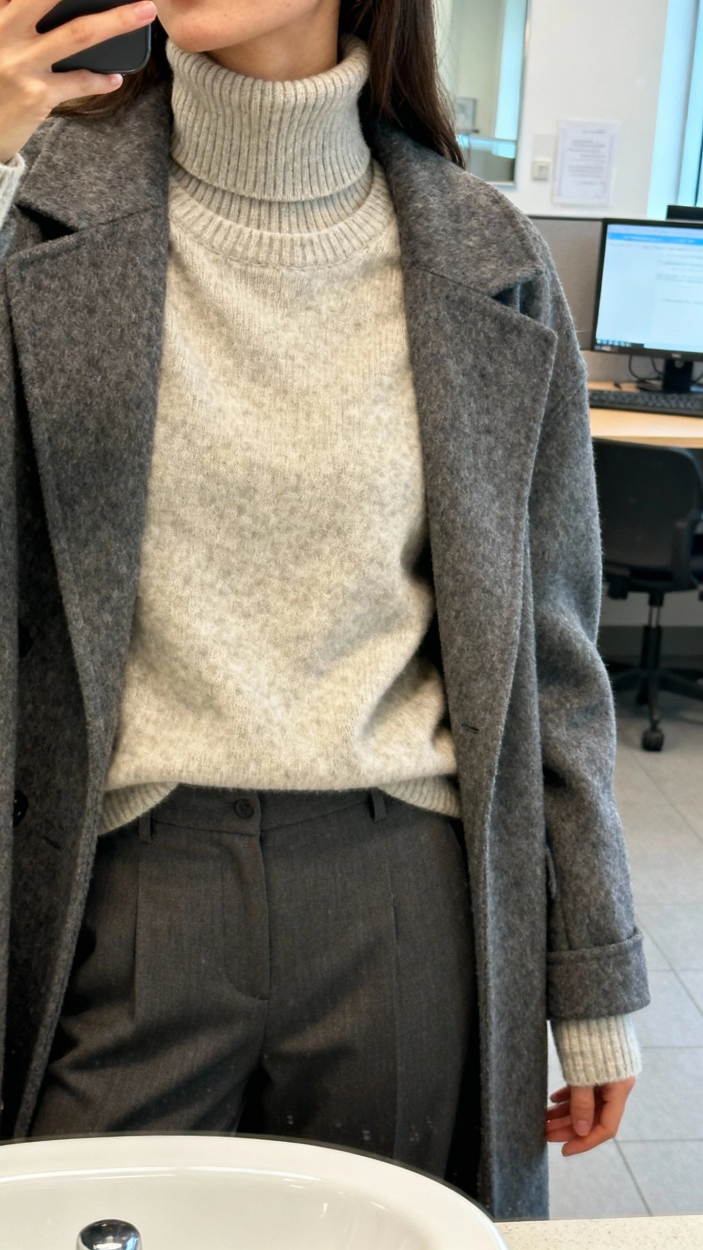 iPhone close crop at neck selfie of a woman showing a layered knit trick: slim turtleneck under a crewneck sweater with coat, sleeves extended to cover wrists, tailored pants, face not visible, office restroom mirror, bright indoor light, iPhone photo quality.