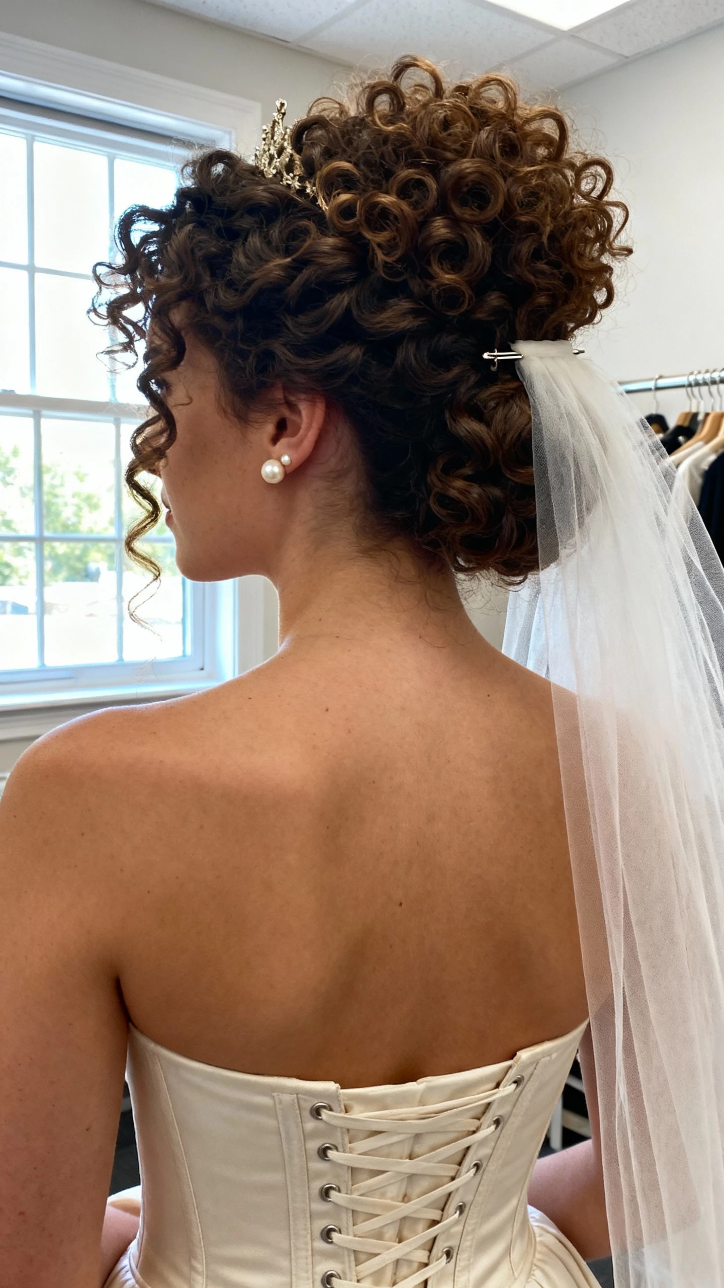 iPhone close crop at neck showing a woman with curly crown volume and defined coils, veil-friendly base pinned at the back, structured corset bodice gown, and pearl studs; face not visible, bridal suite by window, natural daylight, iPhone photo quality.