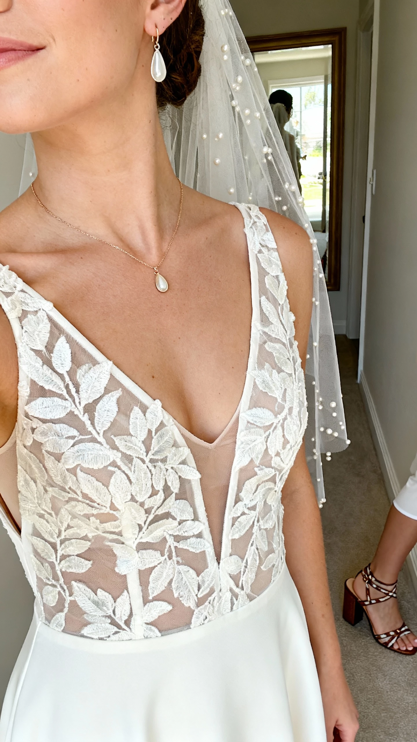 iPhone close crop at shoulders of a woman in a fit-and-flare gown with a plunging illusion V neckline and V back, leafy botanical lace over a crepe base, fingertip veil with scattered pearls, strappy block-heel sandals, delicate lariat necklace, teardrop earrings, seamless shaping shorts; hallway mirror, face not visible, natural daylight, natural iPhone photo quality.