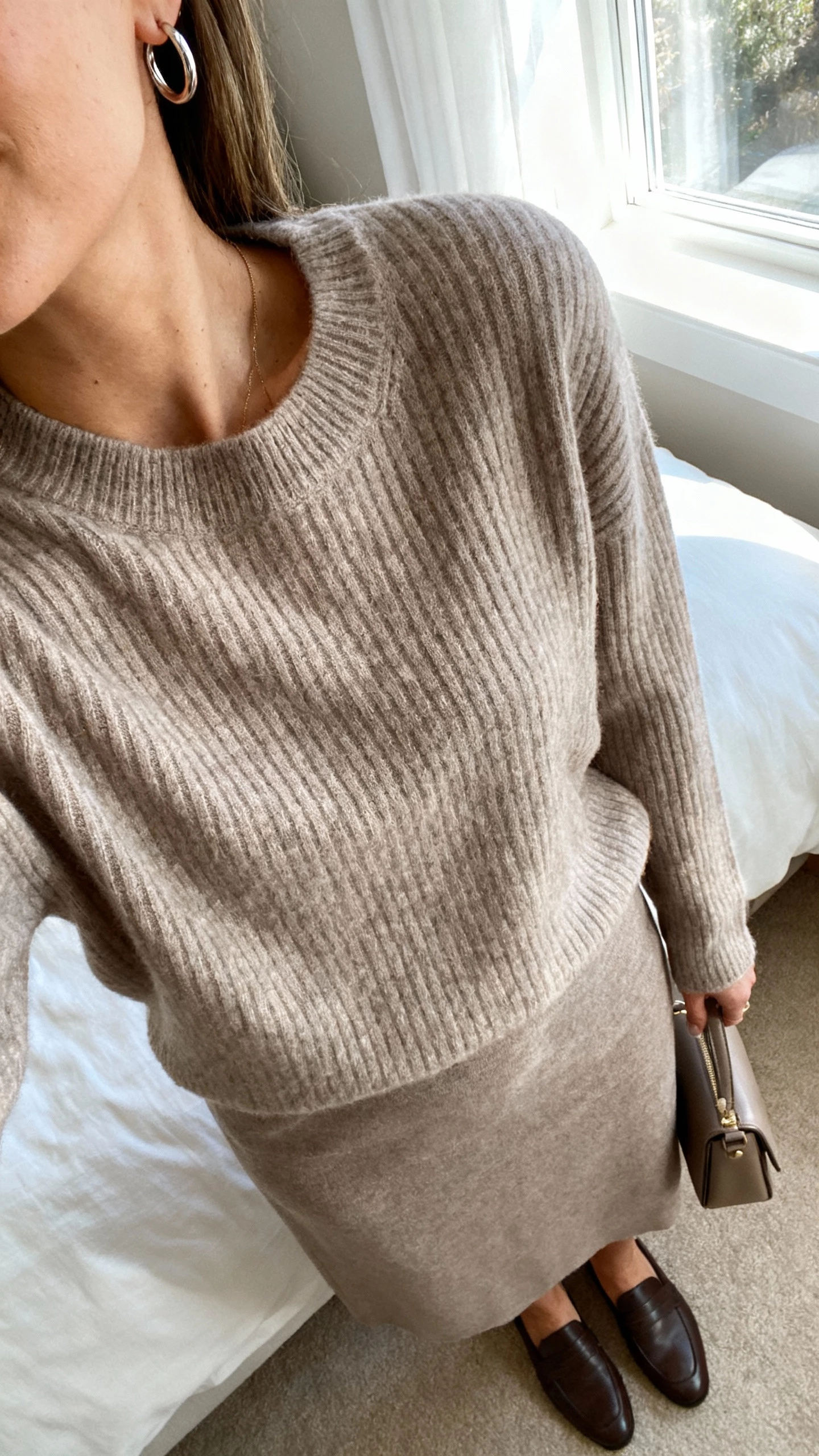 iPhone close crop at shoulders of a woman wearing a matching cashmere-like knit set (ribbed pullover and midi skirt) in soft taupe with delicate hoop earrings, loafers, and a structured mini bag, face not visible, cozy bedroom near window light, natural iPhone photo quality.