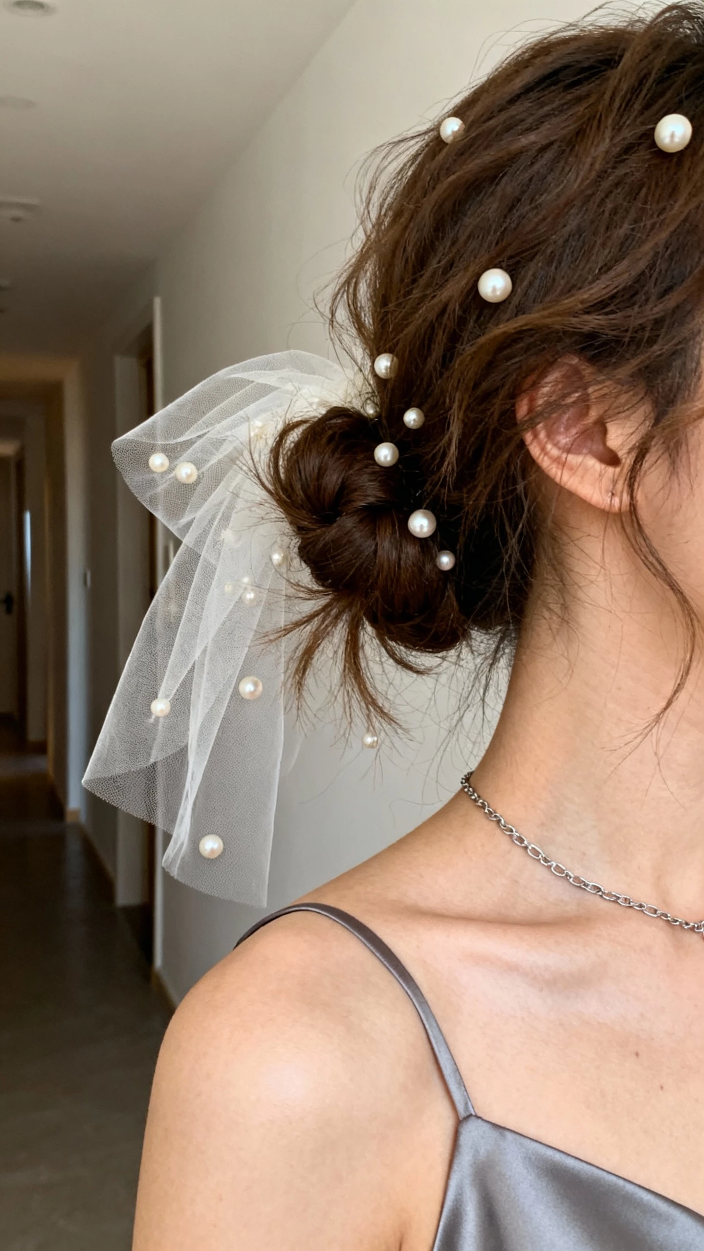 iPhone close crop at shoulders showing a tousled low bun with a delicate face veil and scattered pearls, a sleek slip dress, fine chain necklace, face not visible, softly lit hallway, natural iPhone photo quality.
