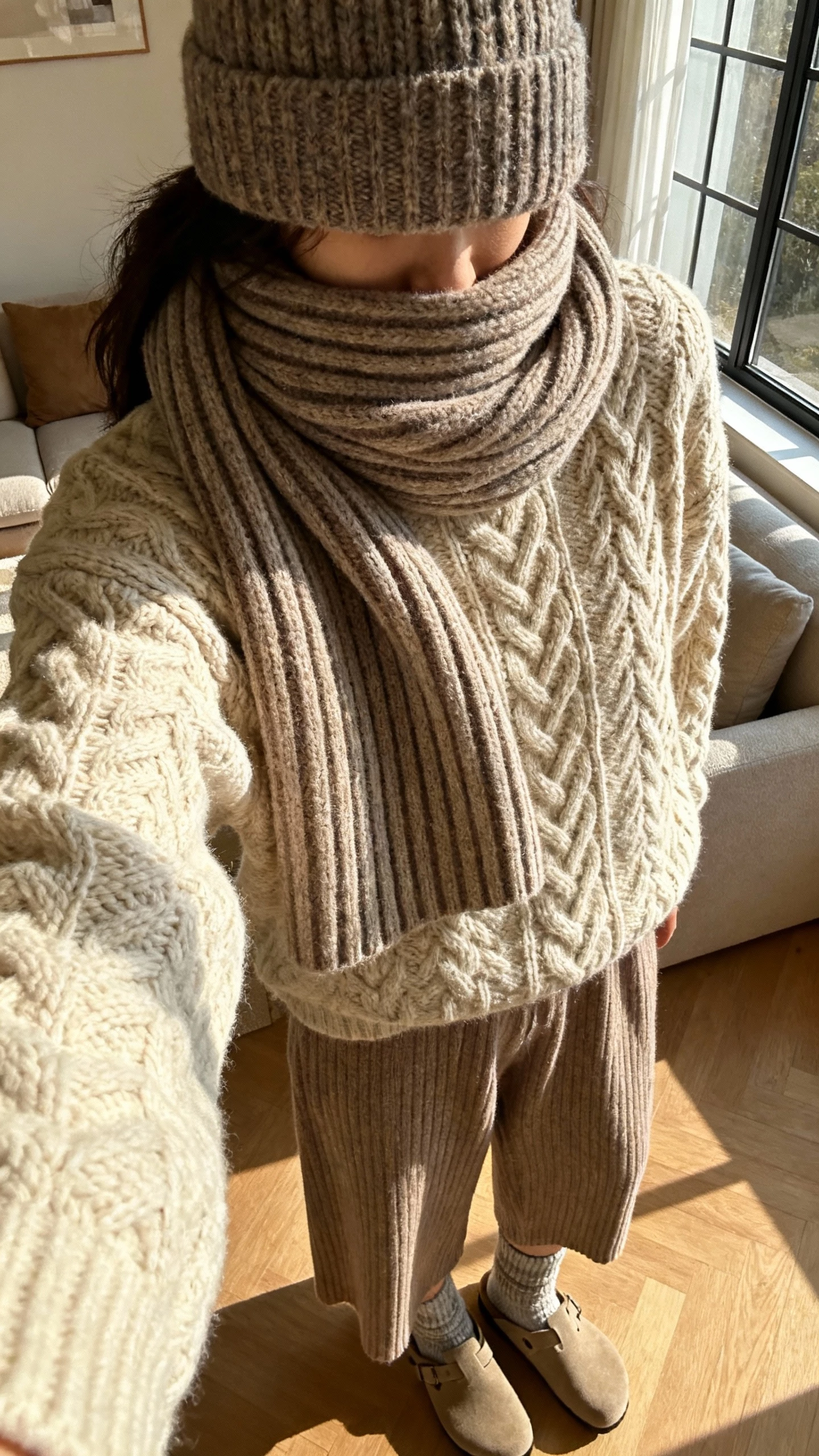 iPhone close-crop selfie at shoulders of a woman layering a cable-knit sweater with a thick ribbed knit scarf and a knit beanie, wide-leg knit pants, wool socks and clogs, face not visible, cozy living room by a window, warm afternoon light, casual iPhone photo.
