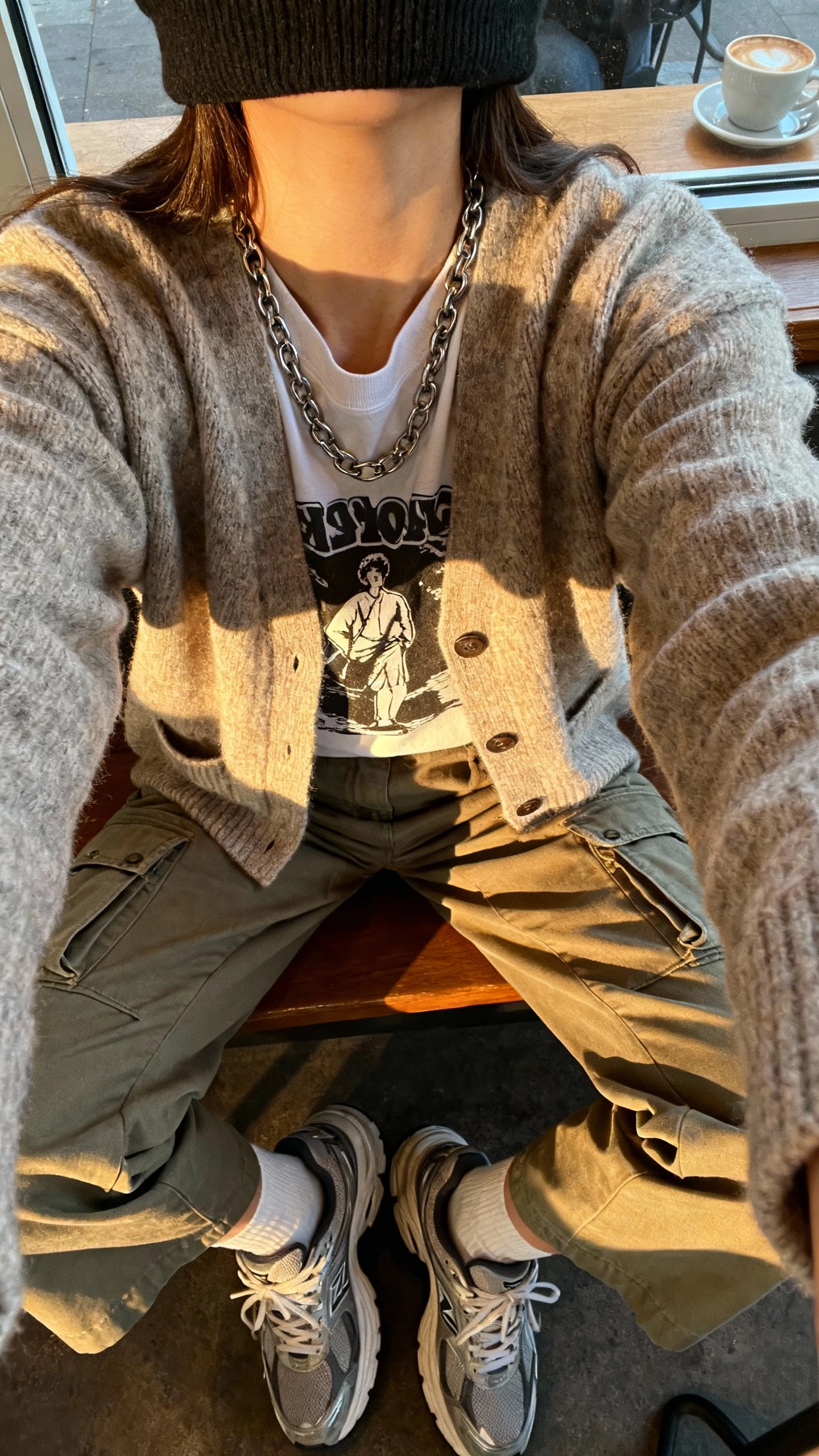 iPhone close-crop selfie at shoulders of a woman wearing a grandpa-style cardigan over a graphic tee, cargo pants, chunky sneakers, chain necklace, beanie, face not visible, coffee shop window seat, soft morning light, natural iPhone photo quality