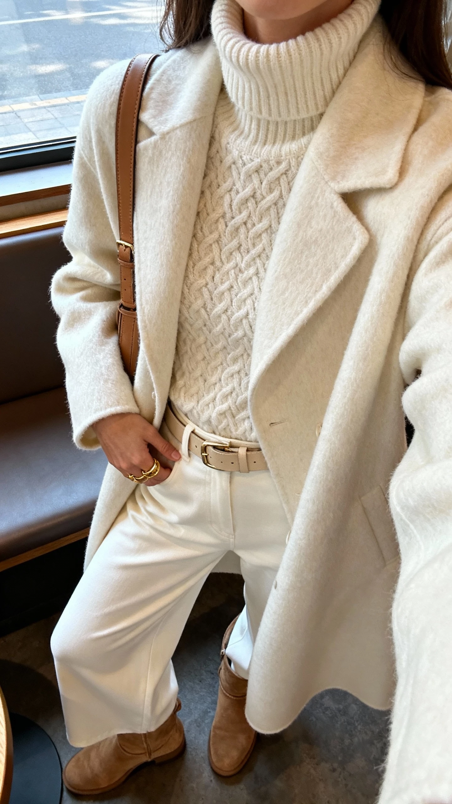 iPhone close-crop selfie at shoulders showing a woman in a luxe monochrome look: cream textured turtleneck, matching cream wool coat, ivory wide-leg trousers, suede ankle boots, tonal belt and leather shoulder bag, gold rings, face not visible, cafe window seat with soft afternoon light, natural iPhone photo