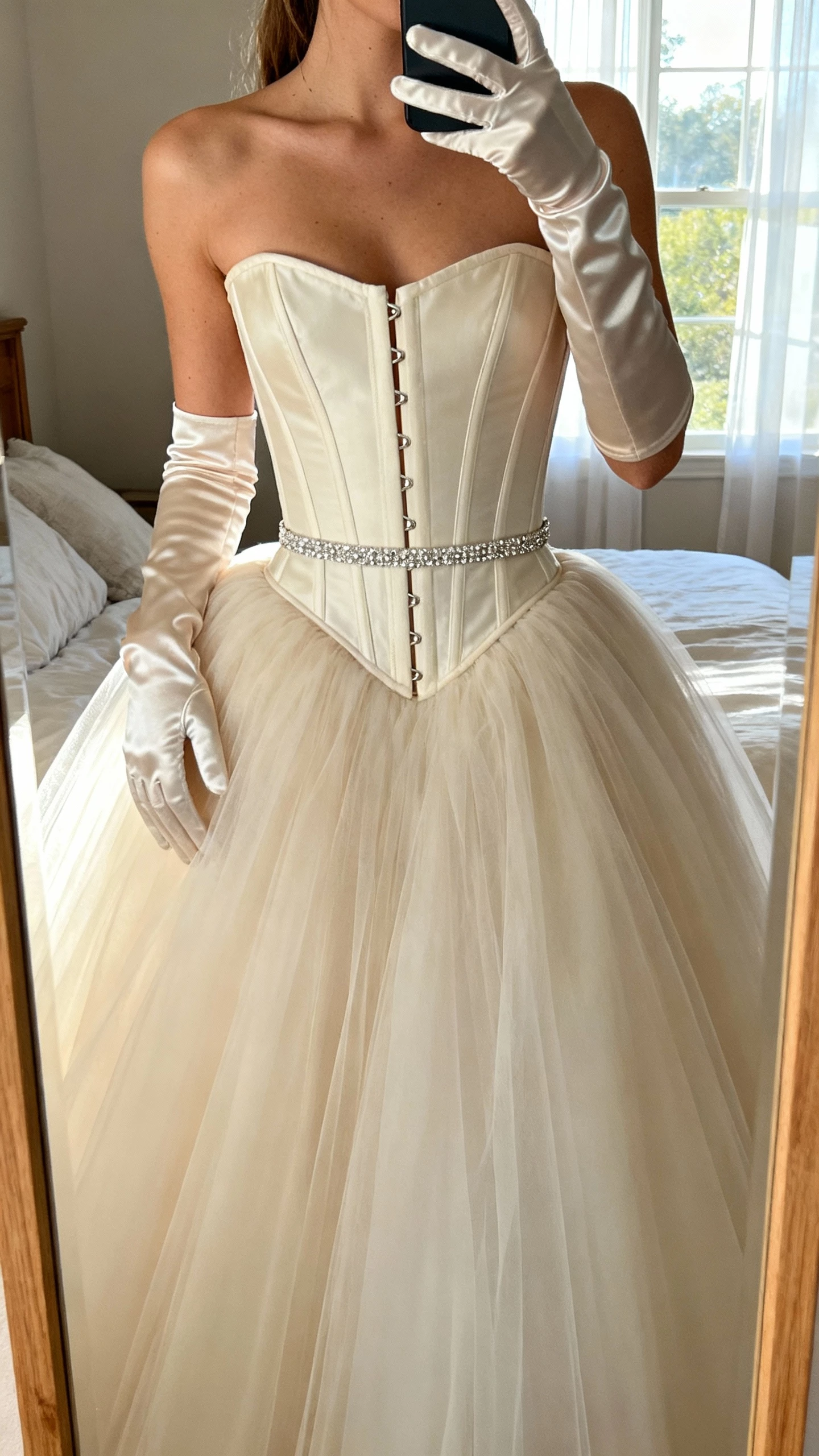 iPhone close-up mirror selfie of a woman wearing a corset couture ball gown with visible boning, cinched waist, full tulle skirt, satin gloves, and a slim crystal belt, face not visible, cropped at shoulders, bedroom mirror, morning window light, natural iPhone photo