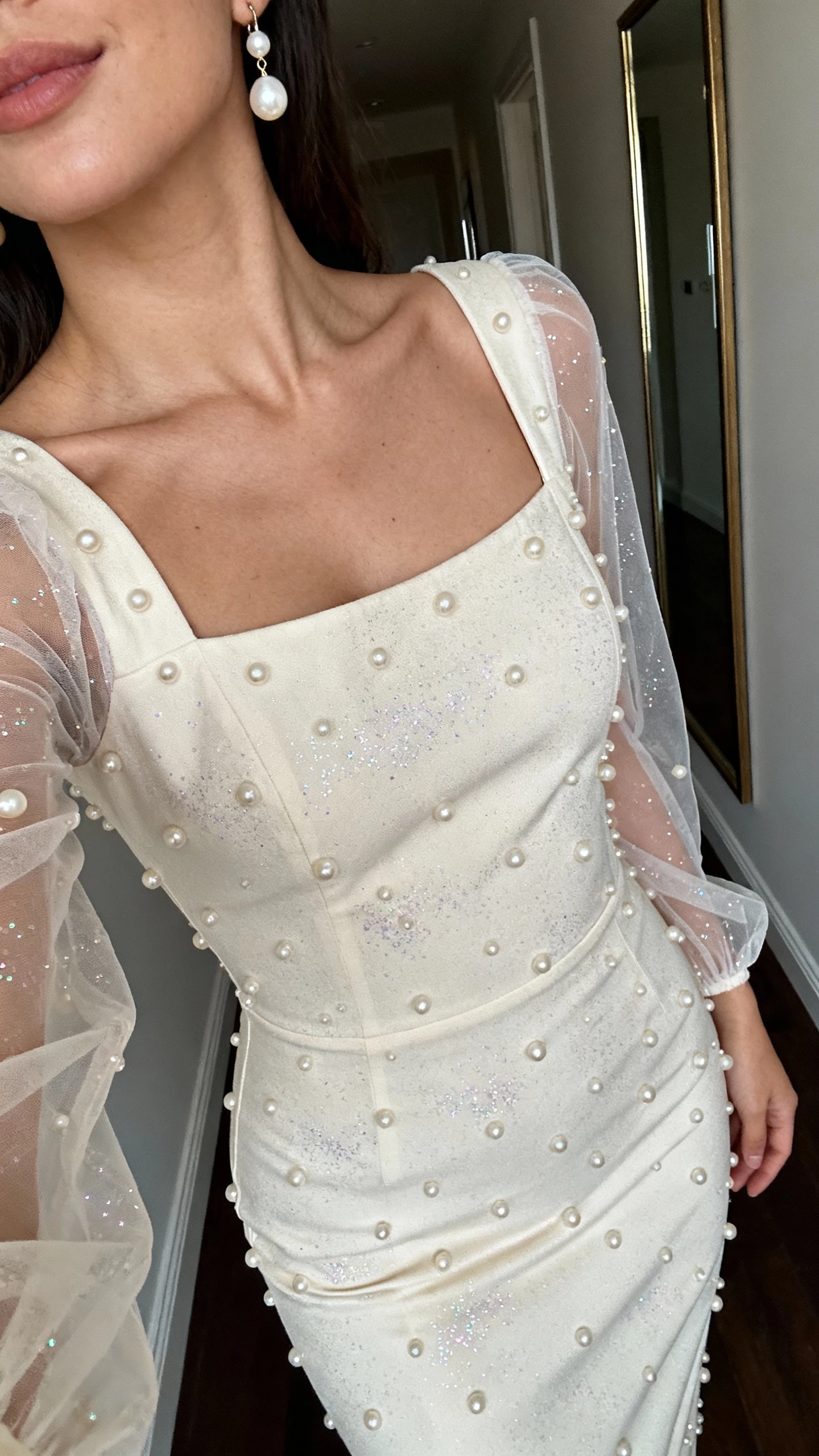 iPhone close-up selfie cropped at neck of a woman wearing a pearl-studded sheath dress with sheer sleeves and subtle sparkle, delicate pearl drop earrings, face not visible, hallway mirror, evening indoor lighting, natural iPhone aesthetic