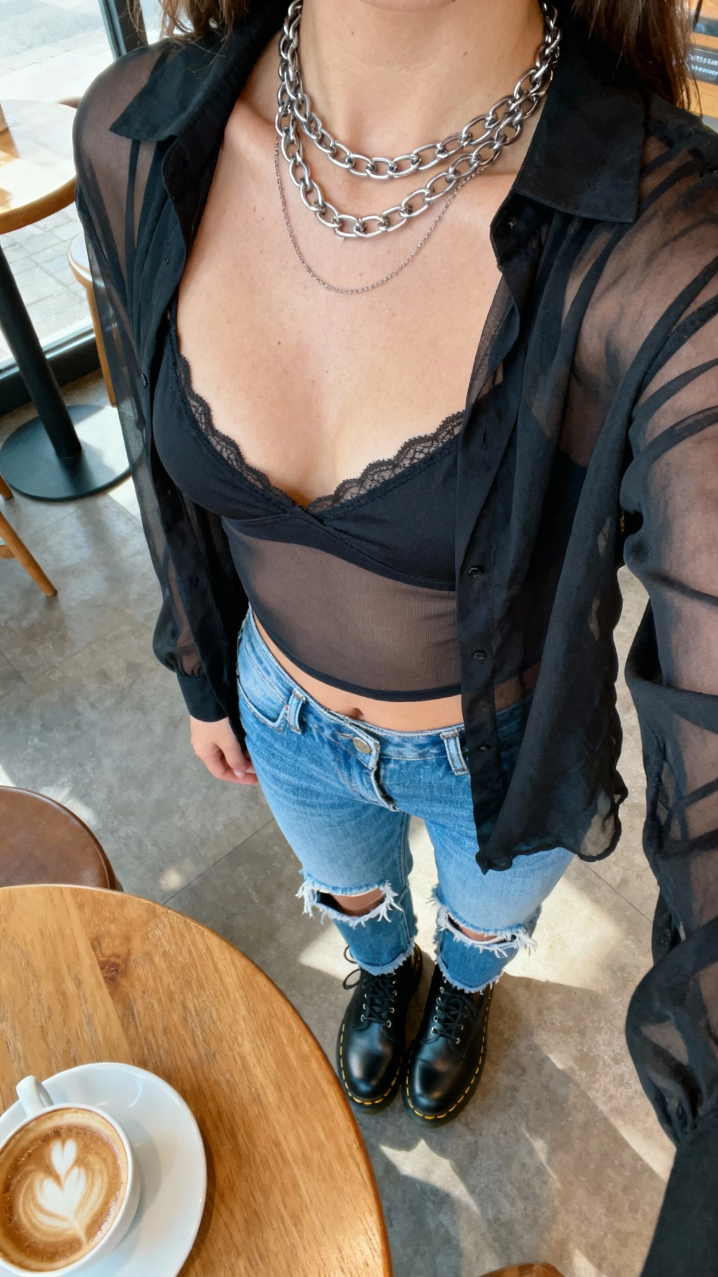 iPhone close-up selfie cropped at shoulders of a woman wearing a black sheer top over a bralette, ripped blue denim, black Doc Martens, layered chain necklace, face not visible, taken at a cozy coffee shop table, soft morning light, natural iPhone photo quality.