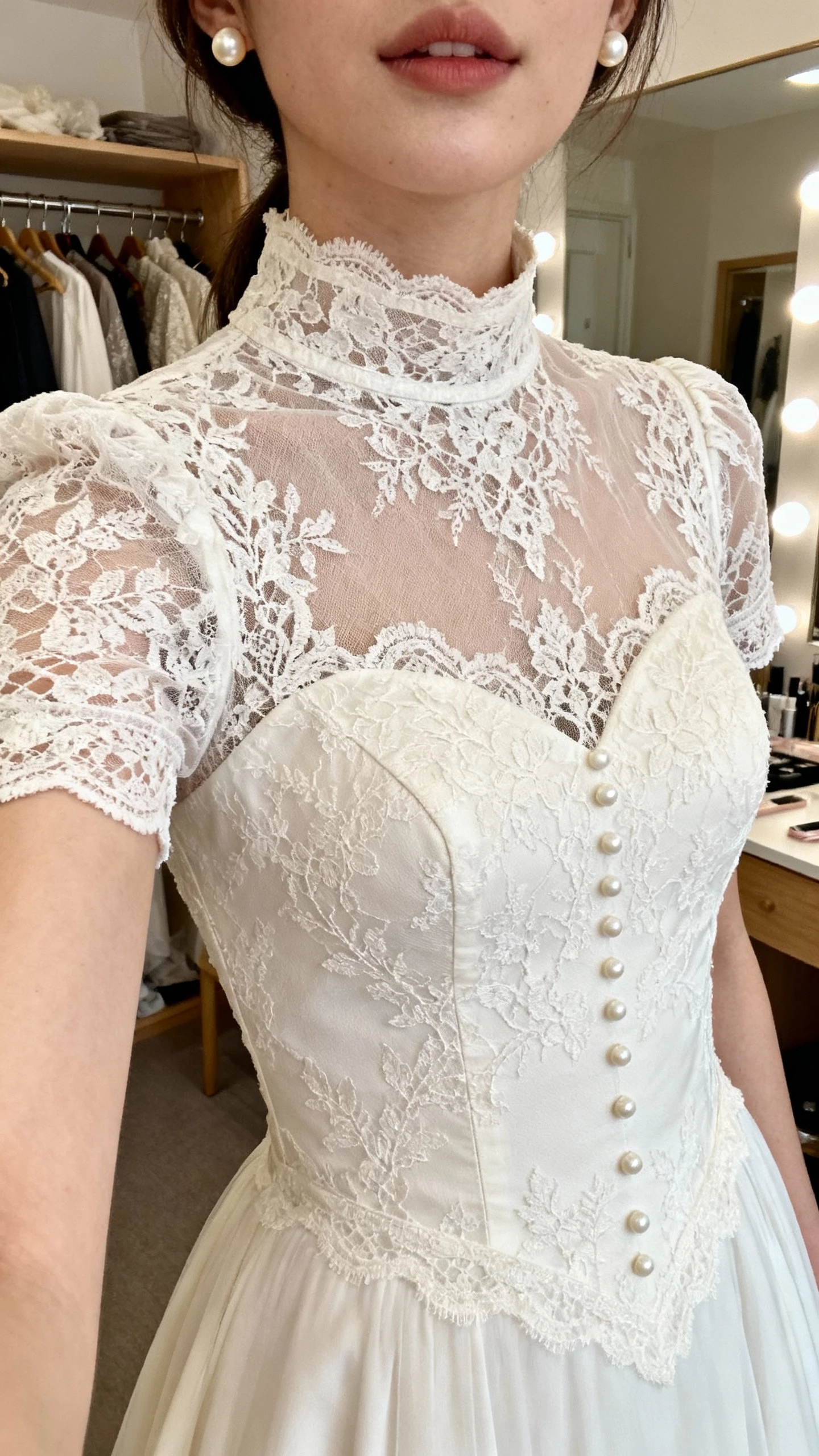 iPhone close-up selfie cropped at shoulders showing a woman wearing a vintage-inspired lace sheath wedding dress with high neck and cap sleeves, pearl studs, face not visible, softly lit dressing room, natural iPhone photo quality.