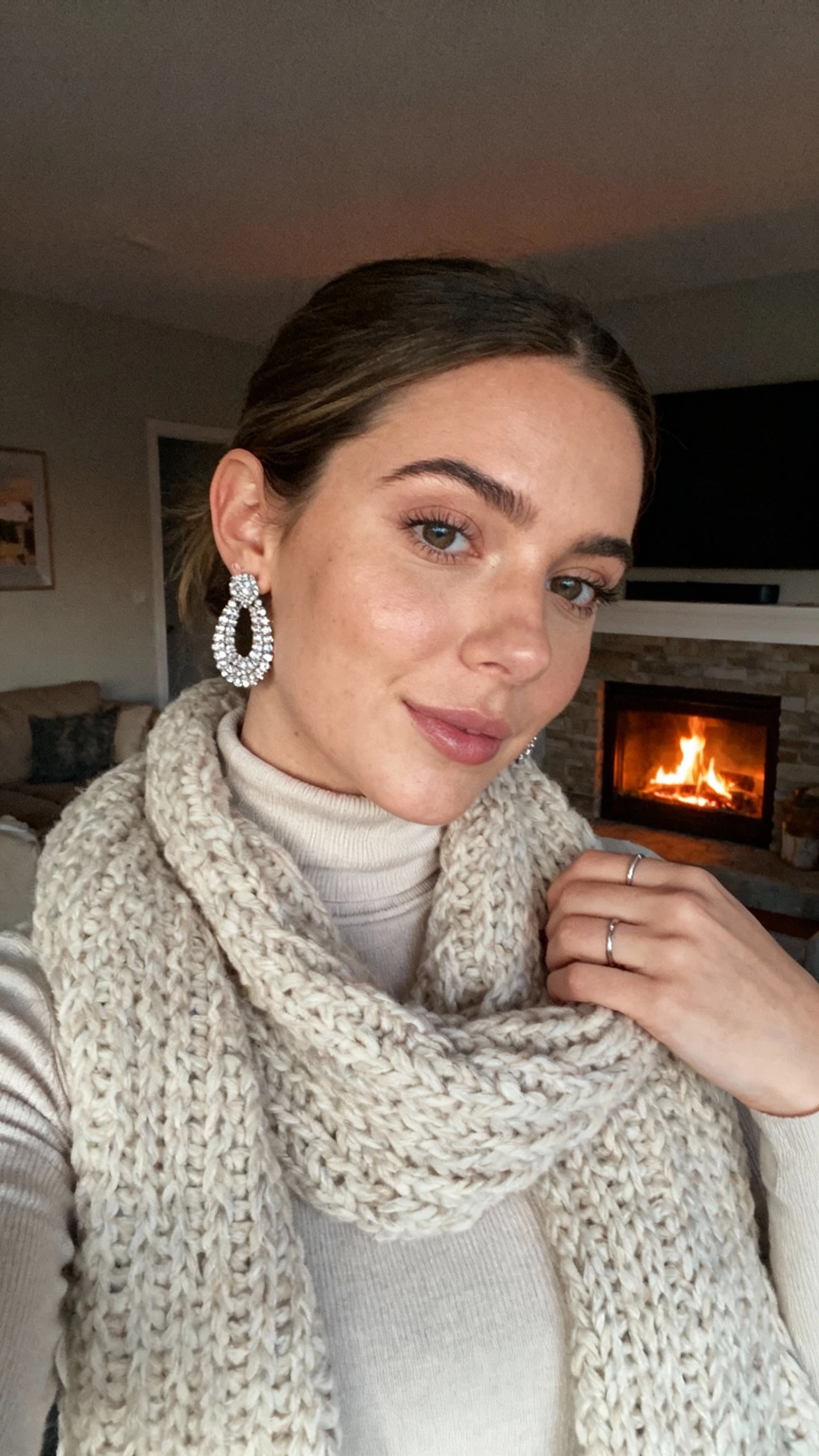 iPhone close-up selfie cropped at shoulders showing a woman wearing sparkly statement earrings, a chunky knit scarf, a fitted turtleneck, and subtle rings, face not visible, fireside living room setting with warm evening light, natural iPhone photo quality.