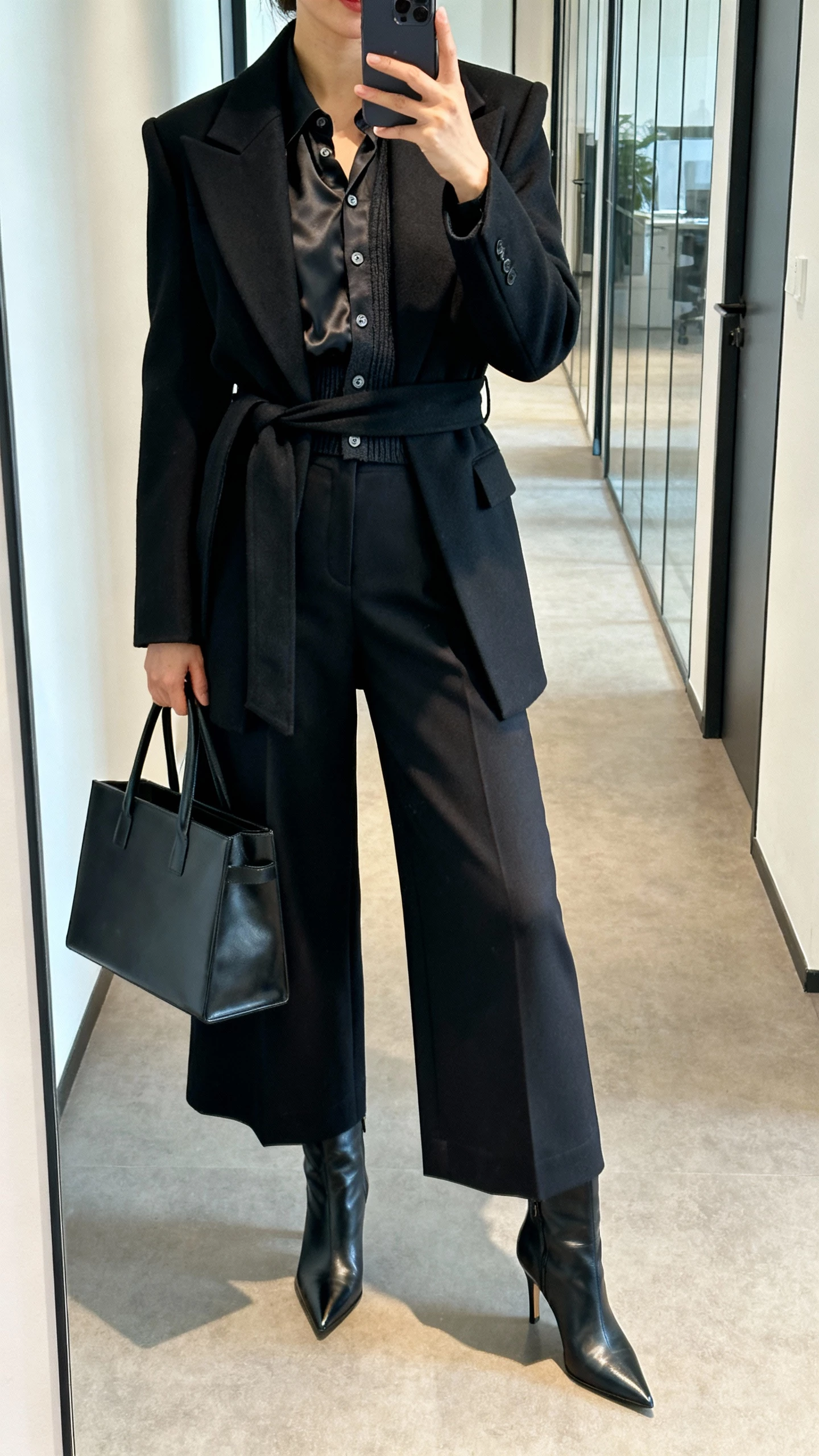 iPhone corridor mirror selfie of a woman in a black midi tailored wrap coat with sharp lapels over a silky button-up blouse and fine cardigan, high-waist wide-leg trousers, pointed heeled boots, boxy leather tote; cropped at shoulders, face not visible, morning office light, casual iPhone photo.