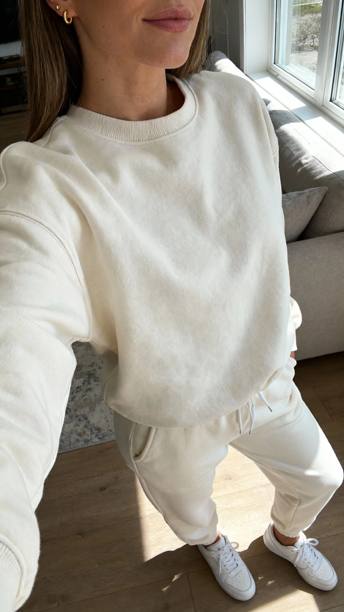 iPhone couch selfie cropped at neck showing a woman in a monochrome jogger set (matching sweatshirt and joggers), sleek white sneakers, minimal gold studs, face not visible, living room with morning window light, natural iPhone photo quality.