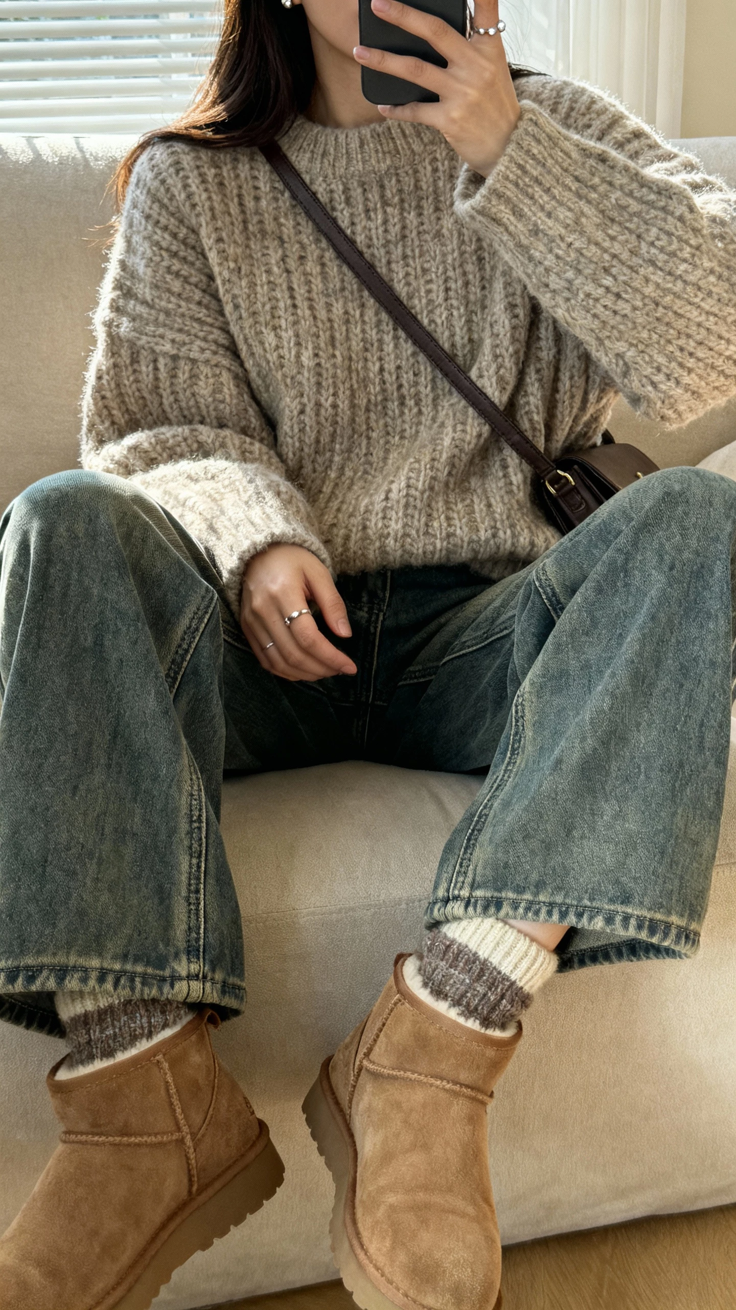 iPhone couch selfie of a woman in an oversized chunky sweater, mid-wash bootcut jeans, comfy ankle boots, cozy socks peeking, simple studs, crossbody bag on side; face not visible, side angle, living room with soft afternoon light, natural iPhone photo quality.