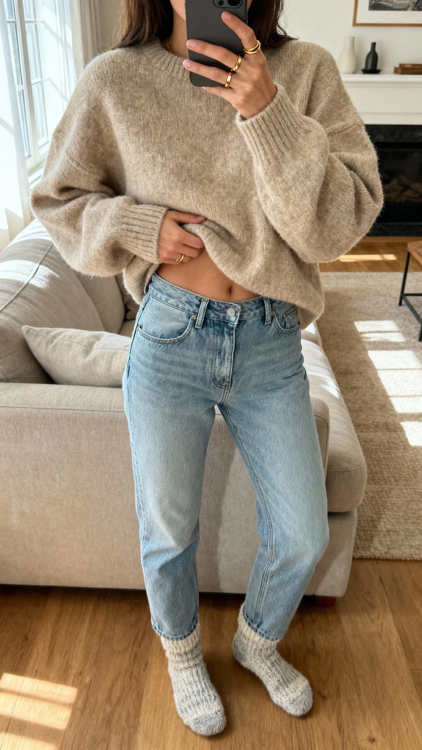 iPhone couch selfie showing a woman in an oversized oatmeal-beige sweater half-tucked into light blue jeans, cozy socks, minimal gold rings, face not visible, living room with soft afternoon window light, natural iPhone photo quality