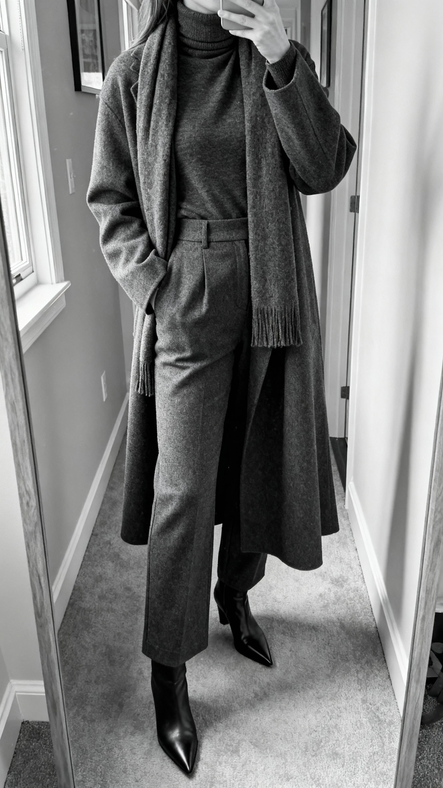 iPhone cropped-at-neck selfie of a woman in a monochrome look: charcoal turtleneck, matching high-waist trousers, long coat in the same tone, pointed boots, and tonal scarf to elongate, face not visible, apartment hallway mirror with morning window light.