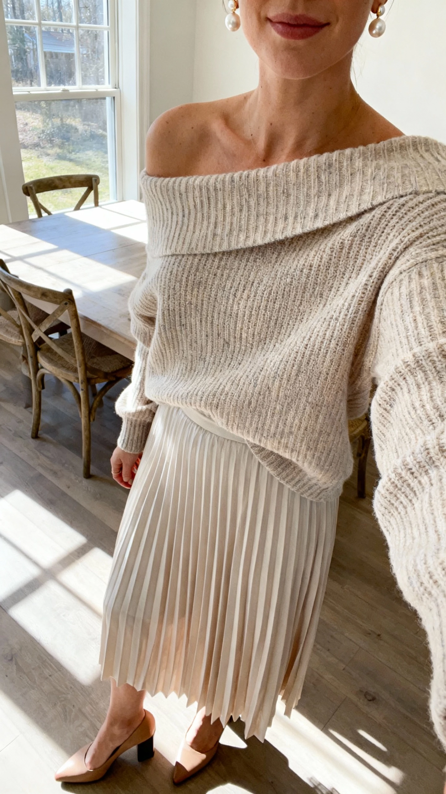 iPhone cropped-at-neck selfie of a woman wearing a pleated midi skirt with an off-shoulder knit sweater, block-heel pumps, and pearl earrings, face not visible, dining room by window, morning light, natural iPhone photo quality
