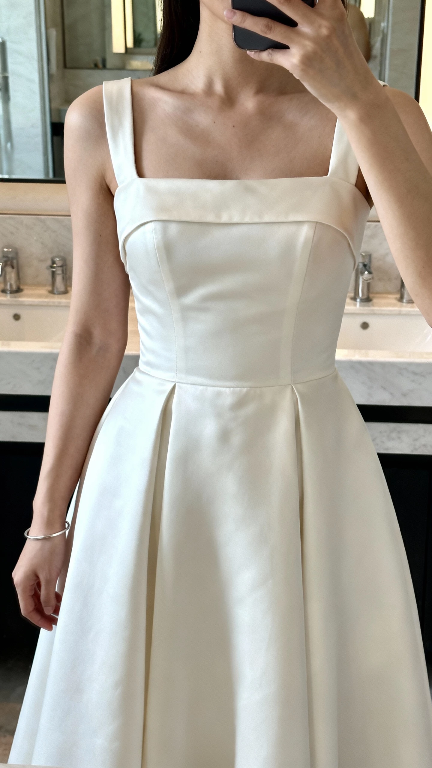 iPhone cropped-at-neck selfie showing a woman in a square-neck A-line wedding dress with structured bodice, wide straps, minimalist satin, and a simple bracelet; face not visible, bathroom mirror with soft daylight, natural iPhone quality.
