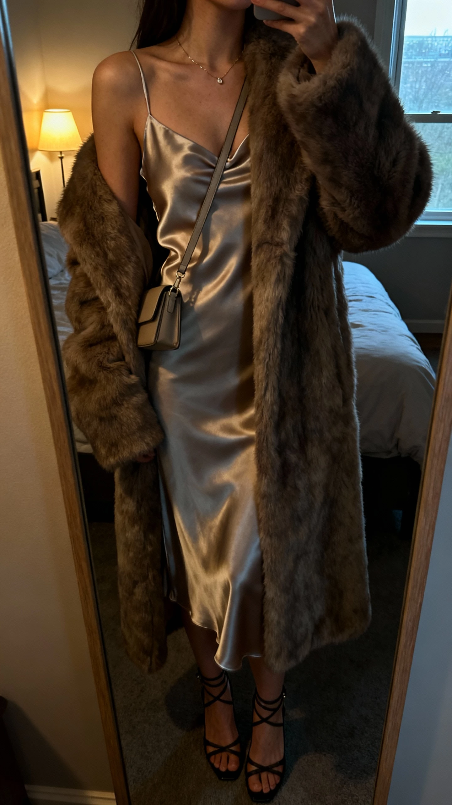 iPhone cropped-at-shoulders mirror selfie of a woman wearing a longline fur coat over a satin slip dress, strappy heels, slim shoulder bag, dainty necklace, face not visible, dim bedroom lamp with window glow, natural iPhone photo quality.