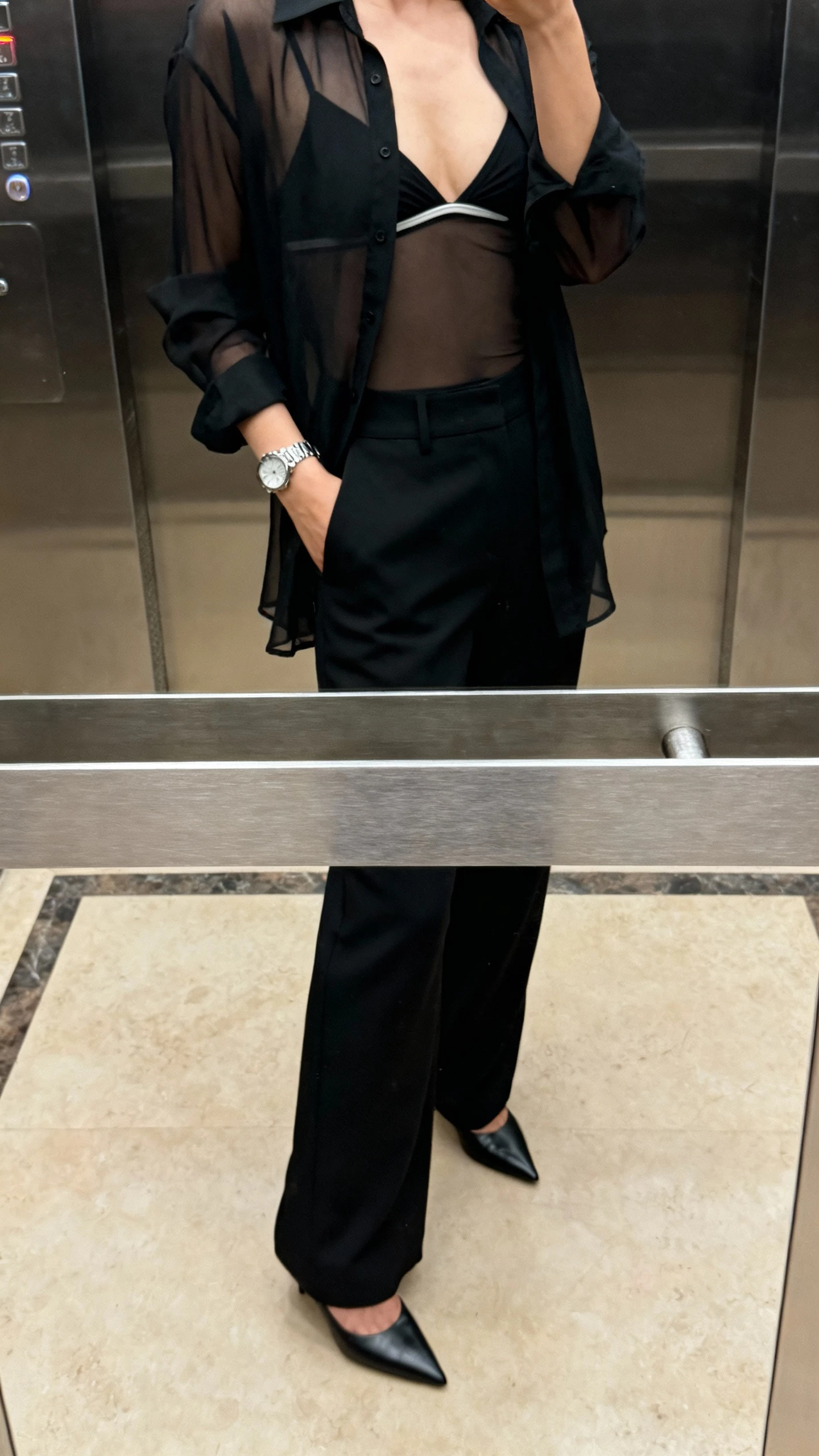 iPhone cropped-at-shoulders selfie of a woman wearing a sheer black top over a bralette with tailored black trousers, pointed heels, and a wristwatch, face not visible, elevator mirror with evening indoor lighting, natural iPhone photo quality