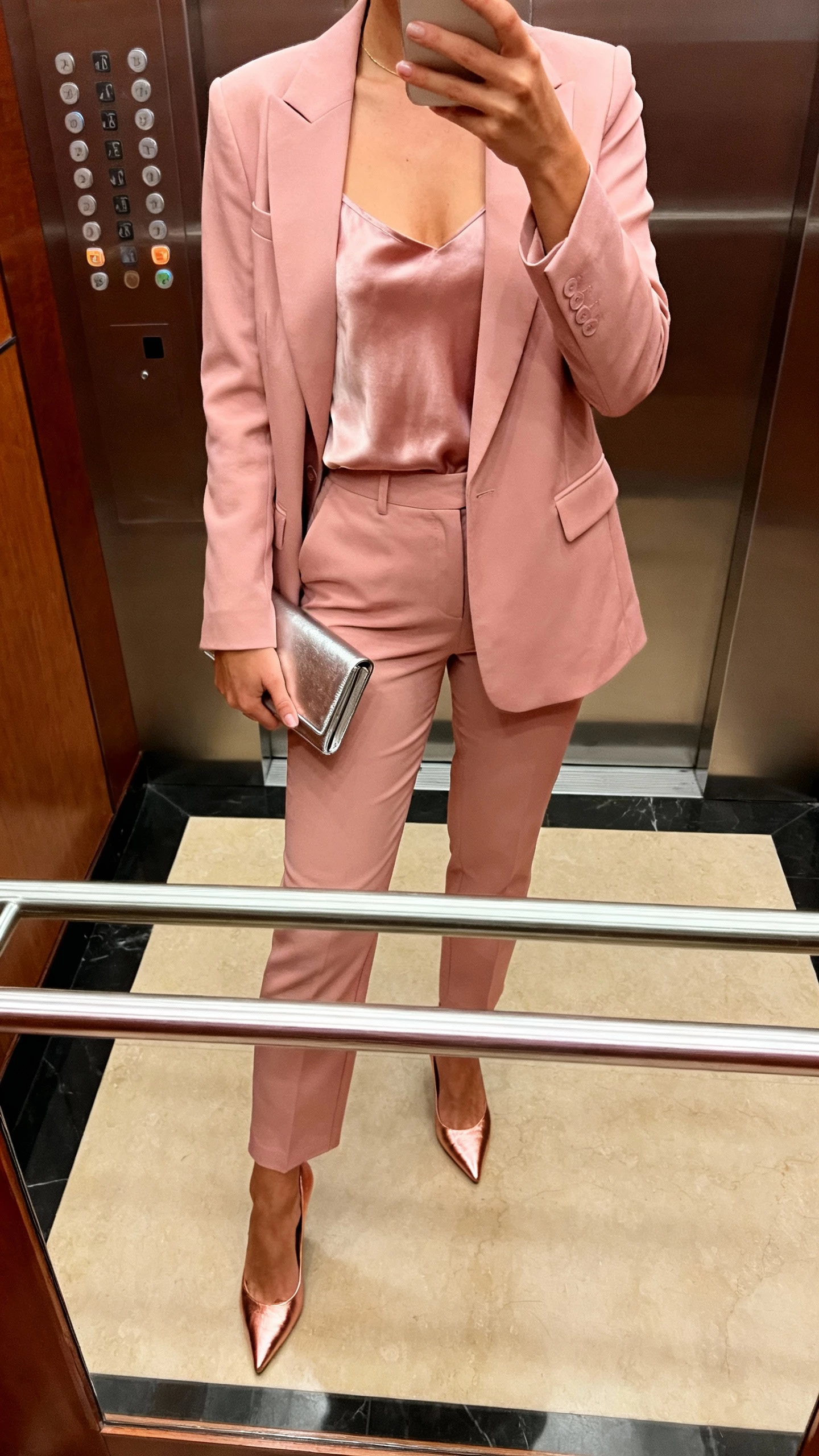 iPhone cropped-at-shoulders selfie showing a woman in a blush-pink suit (blazer and tailored trousers), silky camisole, rose-gold heels, and a slim metallic clutch, face not visible, hotel elevator mirror with warm indoor lighting, natural iPhone quality.
